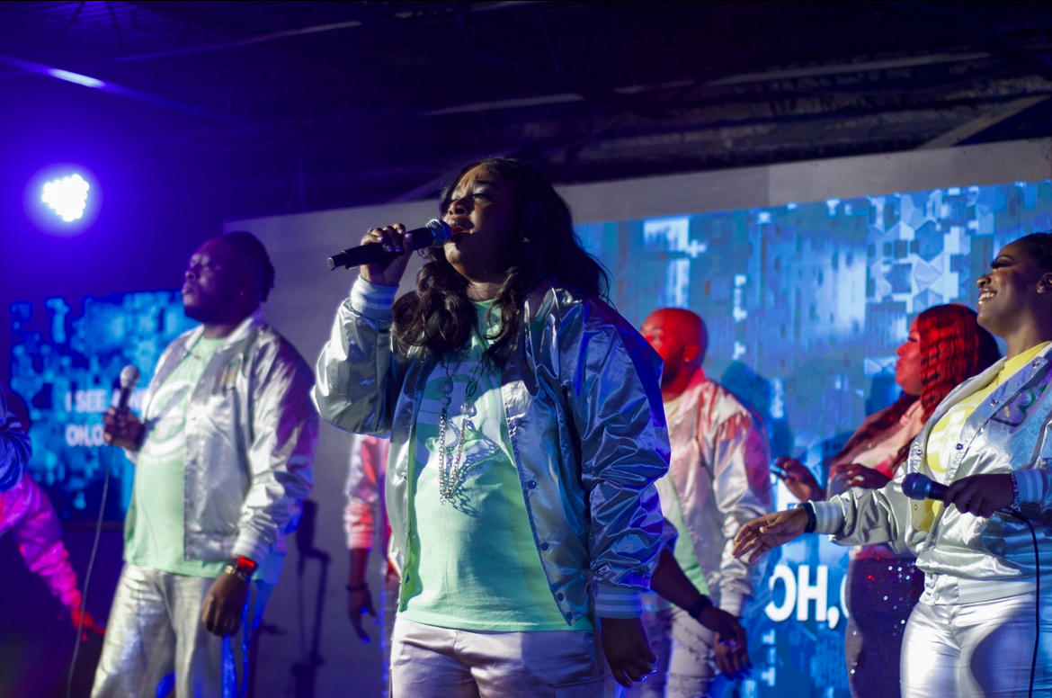 Group of people performing on stage with a woman singing into a microphone, blue background, stage lighting, diverse attire.