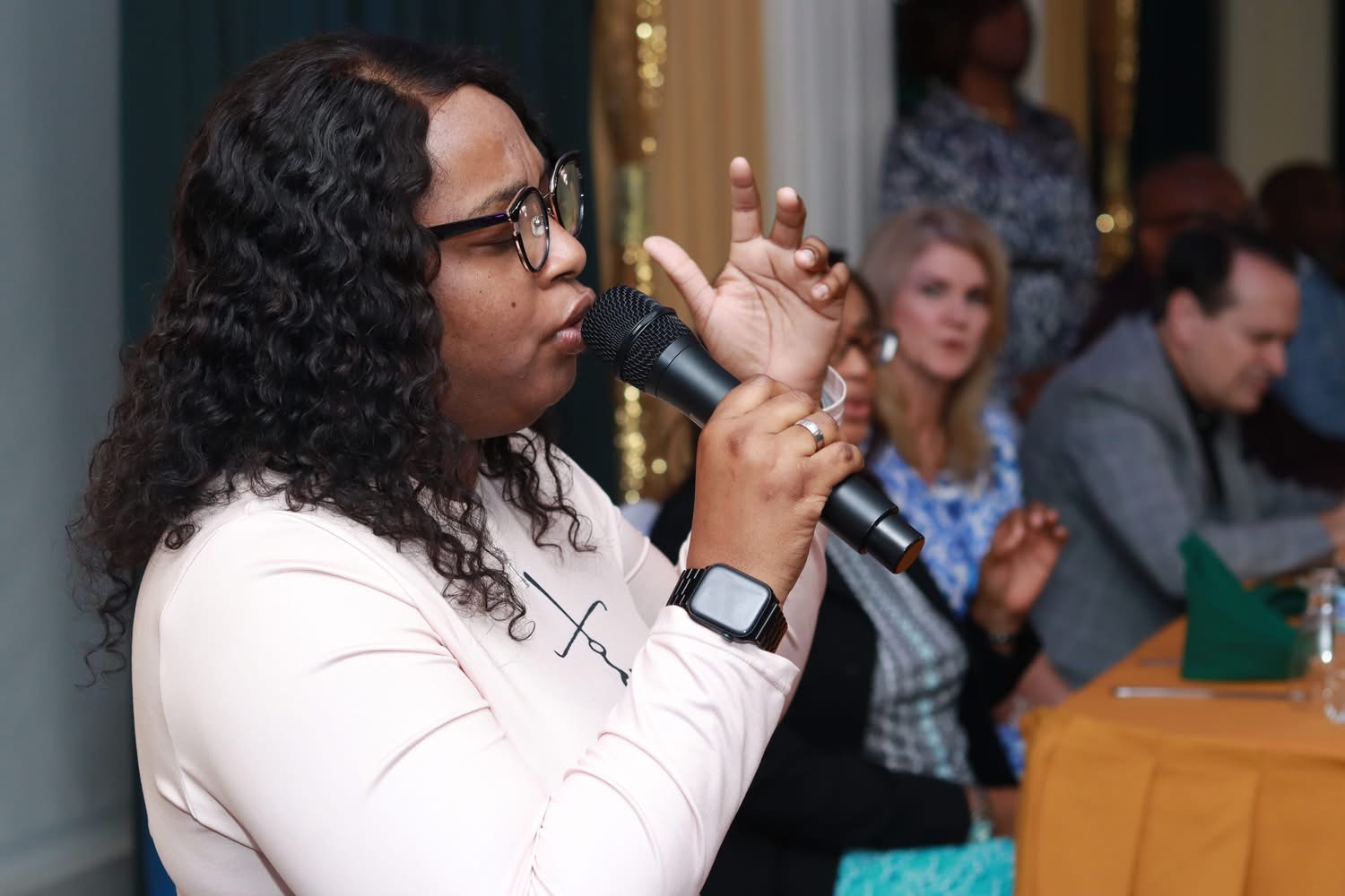 Woman with curly hair and glasses speaking into a microphone at a table with people in the background.