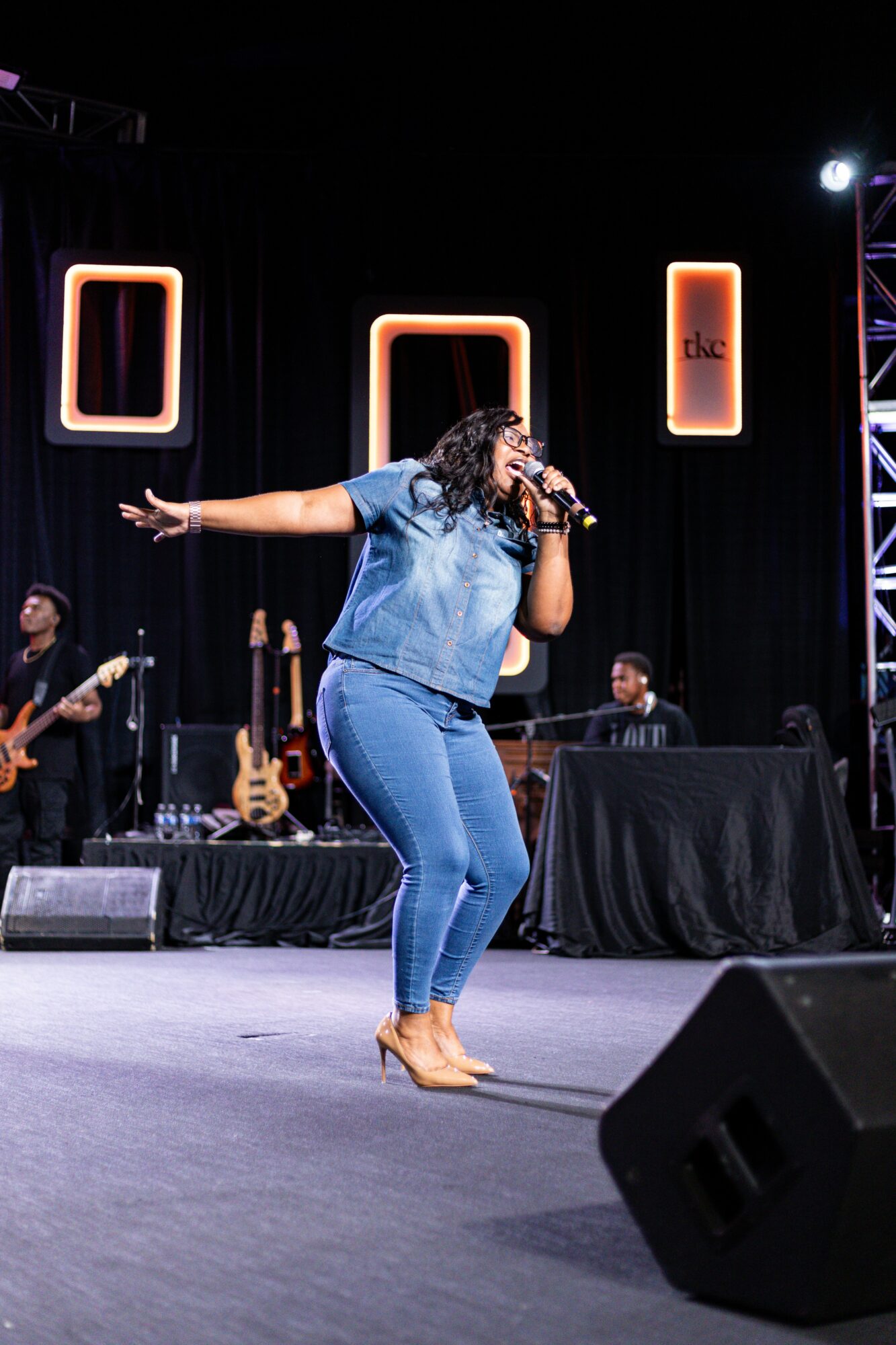 Woman singing on stage with microphone, band members, and musical instruments in background.