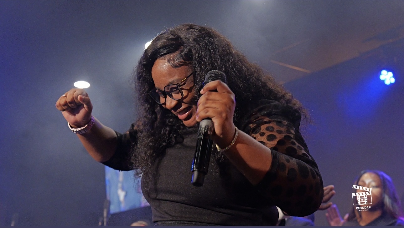 Woman with curly hair and glasses holding a microphone, smiling and raising her fist on stage.