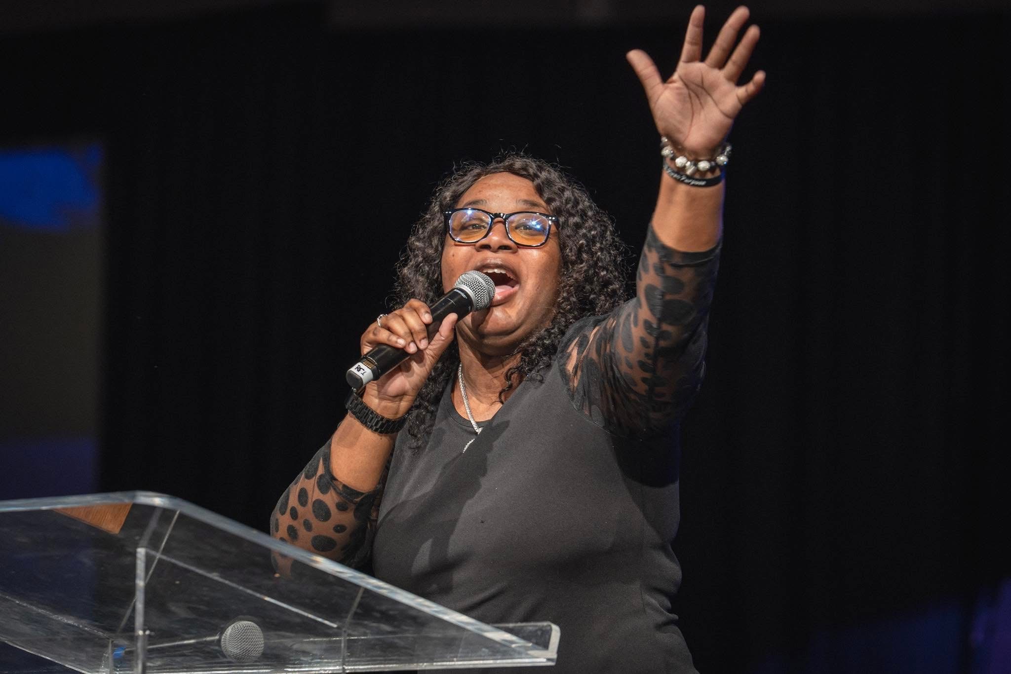 Woman with curly hair and glasses speaking into a microphone, raising her hand, on stage with a black curtain background.