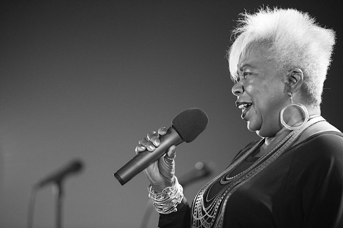 A woman with short, white hair singing into a microphone on stage, with a blurred background.