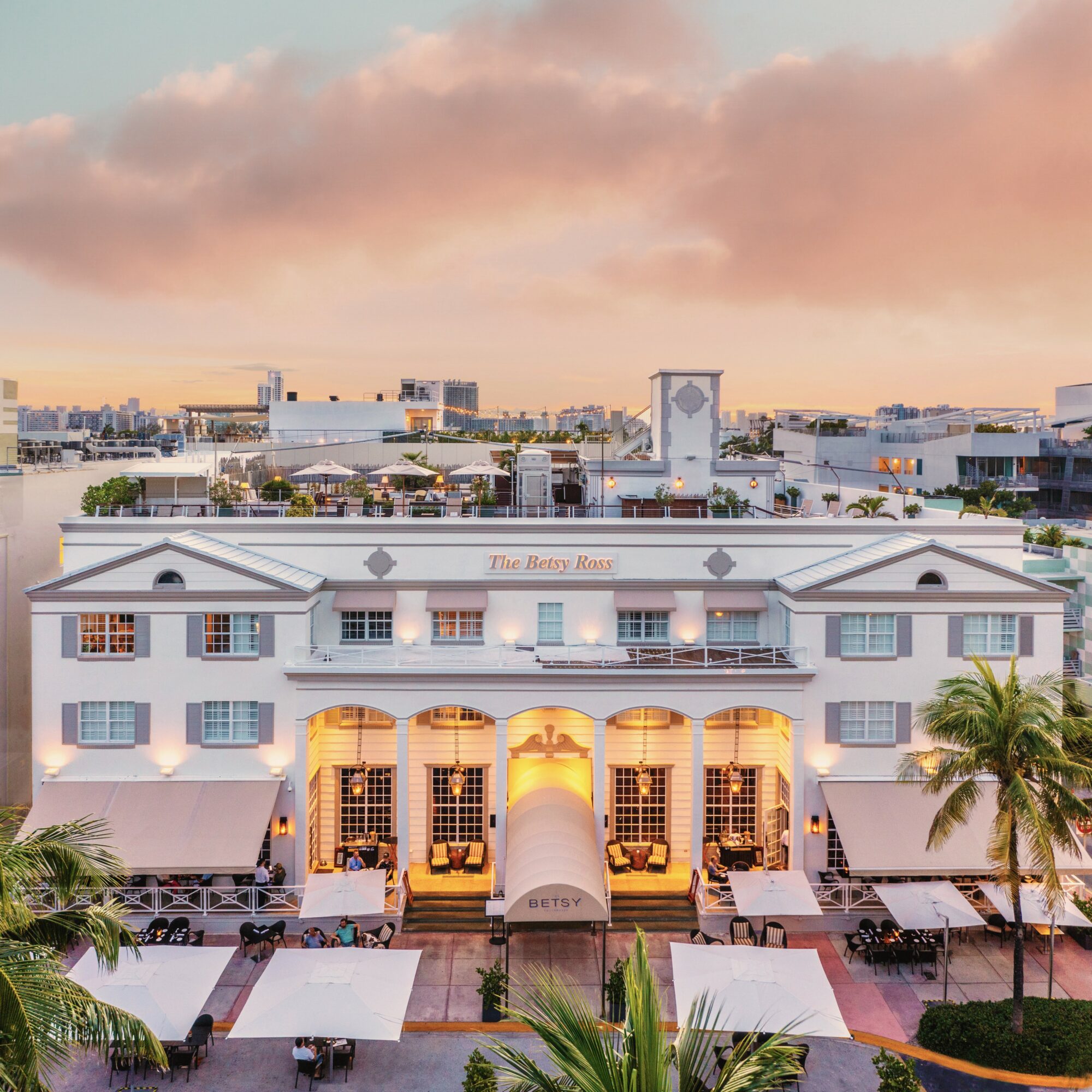White building with arched windows and balconies, palm trees, outdoor seating, and a pink sky in the background.
