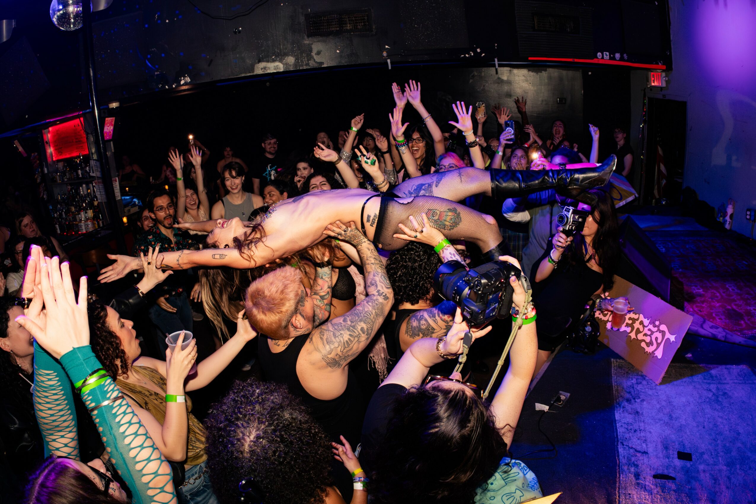 Crowd at nightclub lifting a person horizontally above their heads with hands raised, colorful lighting, dance floor, and DJ booth visible.