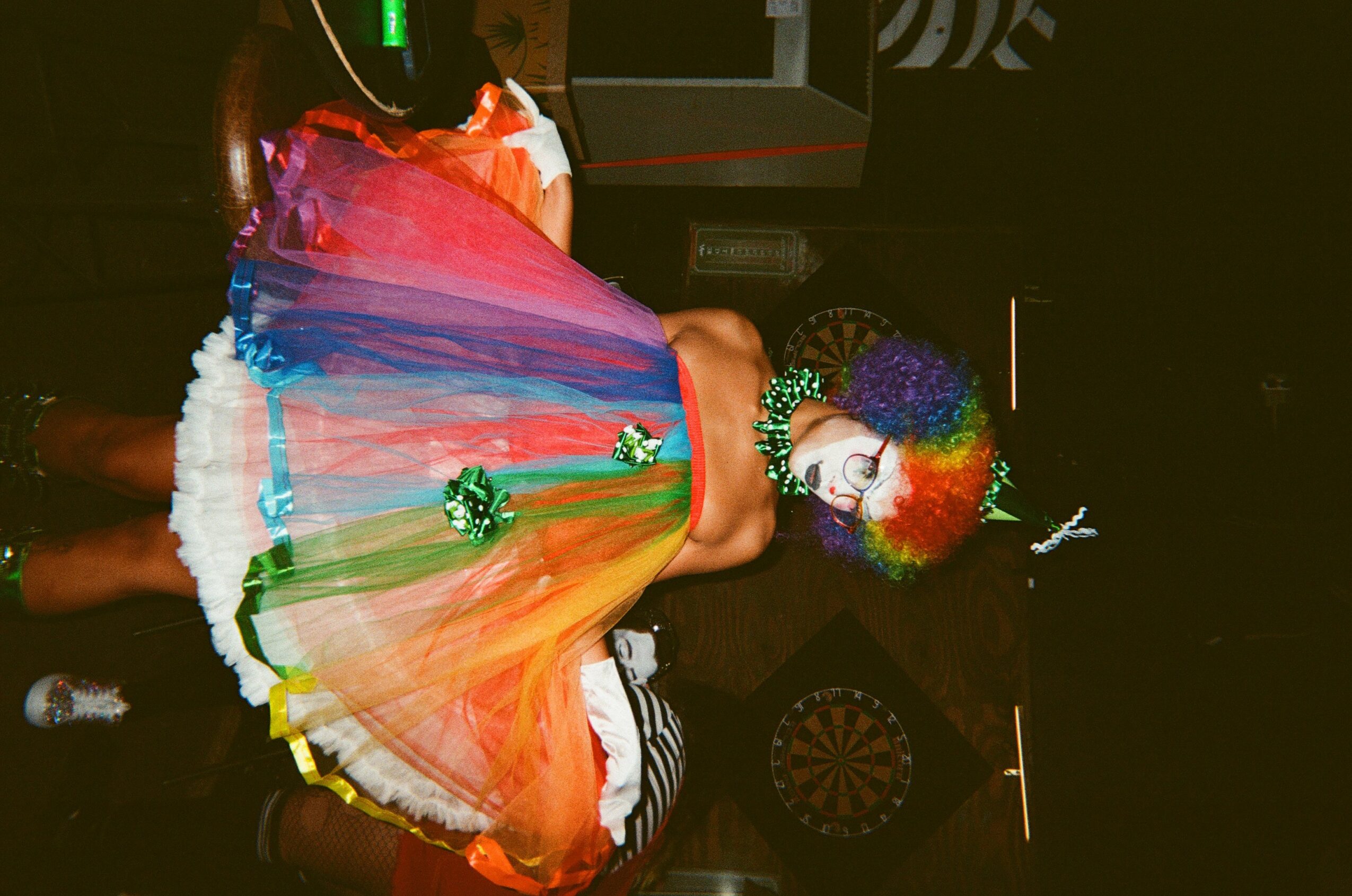 Person in colorful rainbow dress and rainbow wig, holding a rainbow umbrella, standing indoors with dark background.