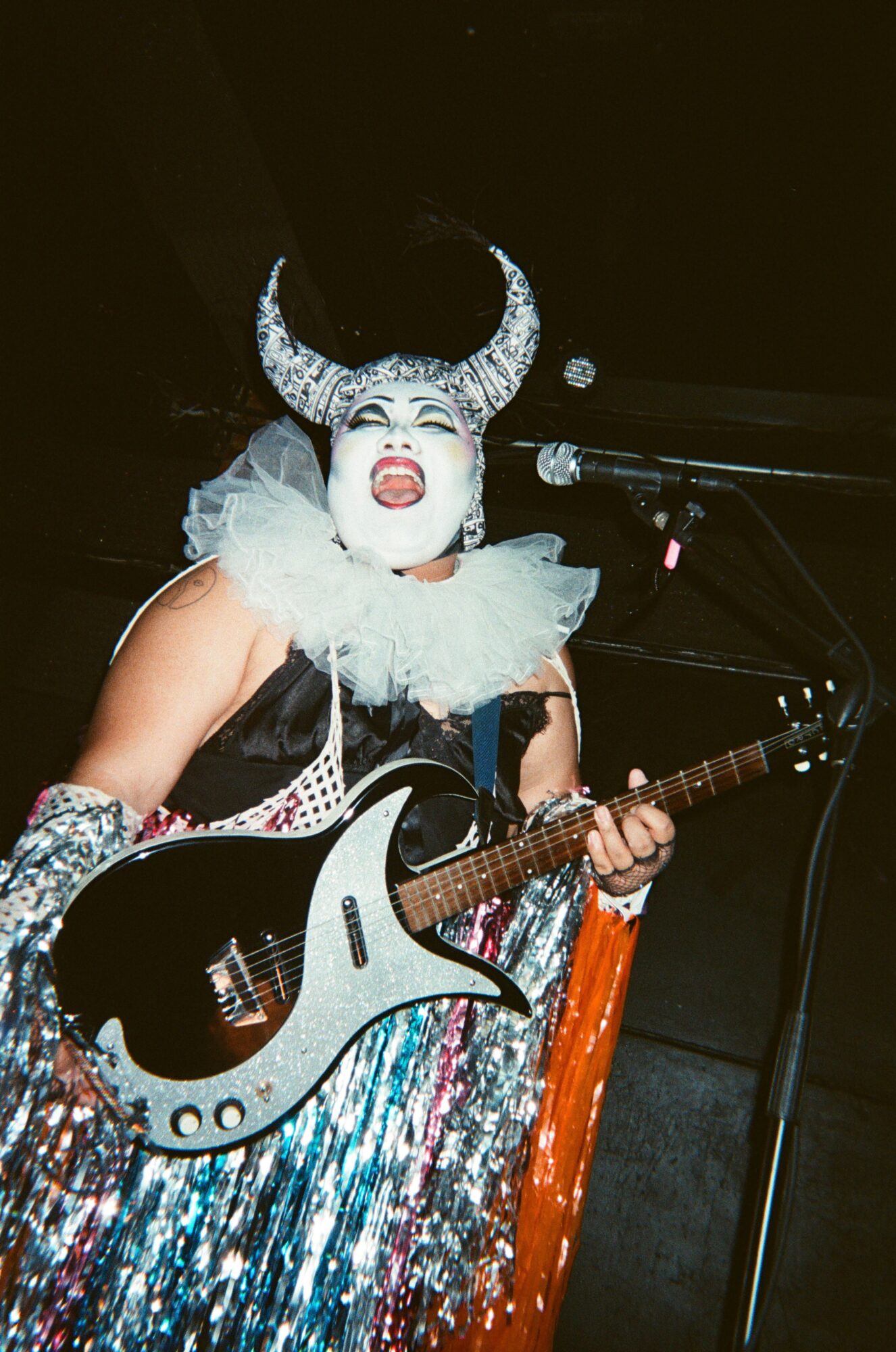 Performer with elaborate horned headpiece, white face makeup, and colorful costume playing guitar on stage.