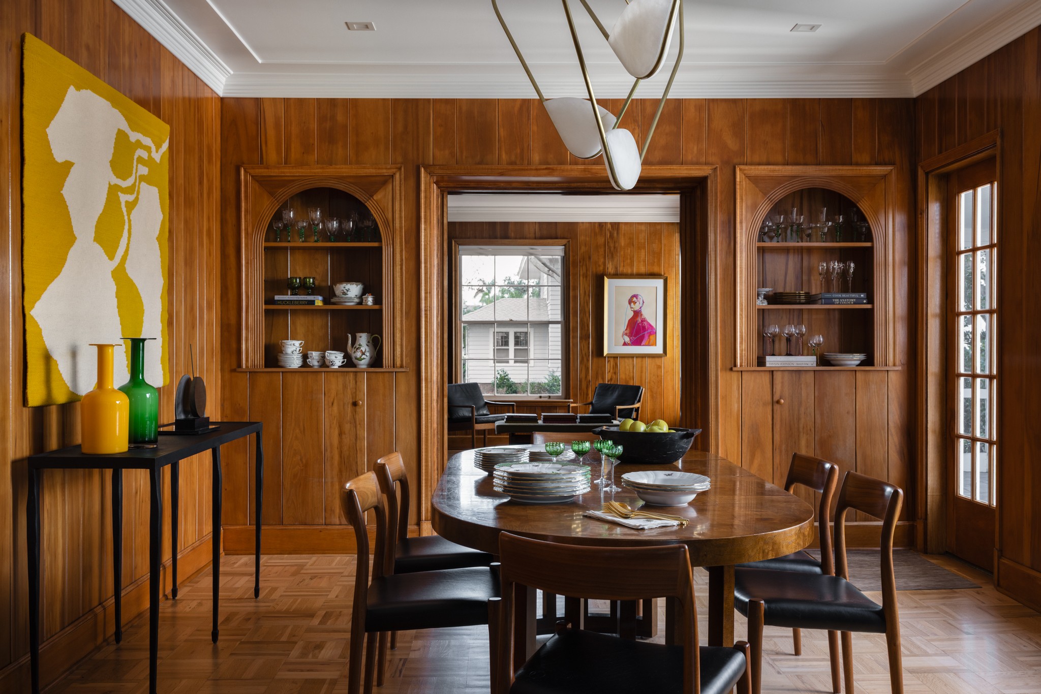 Dining room with wooden paneling, round table, four chairs, and built-in shelves, with artwork on the walls and a chandelier.