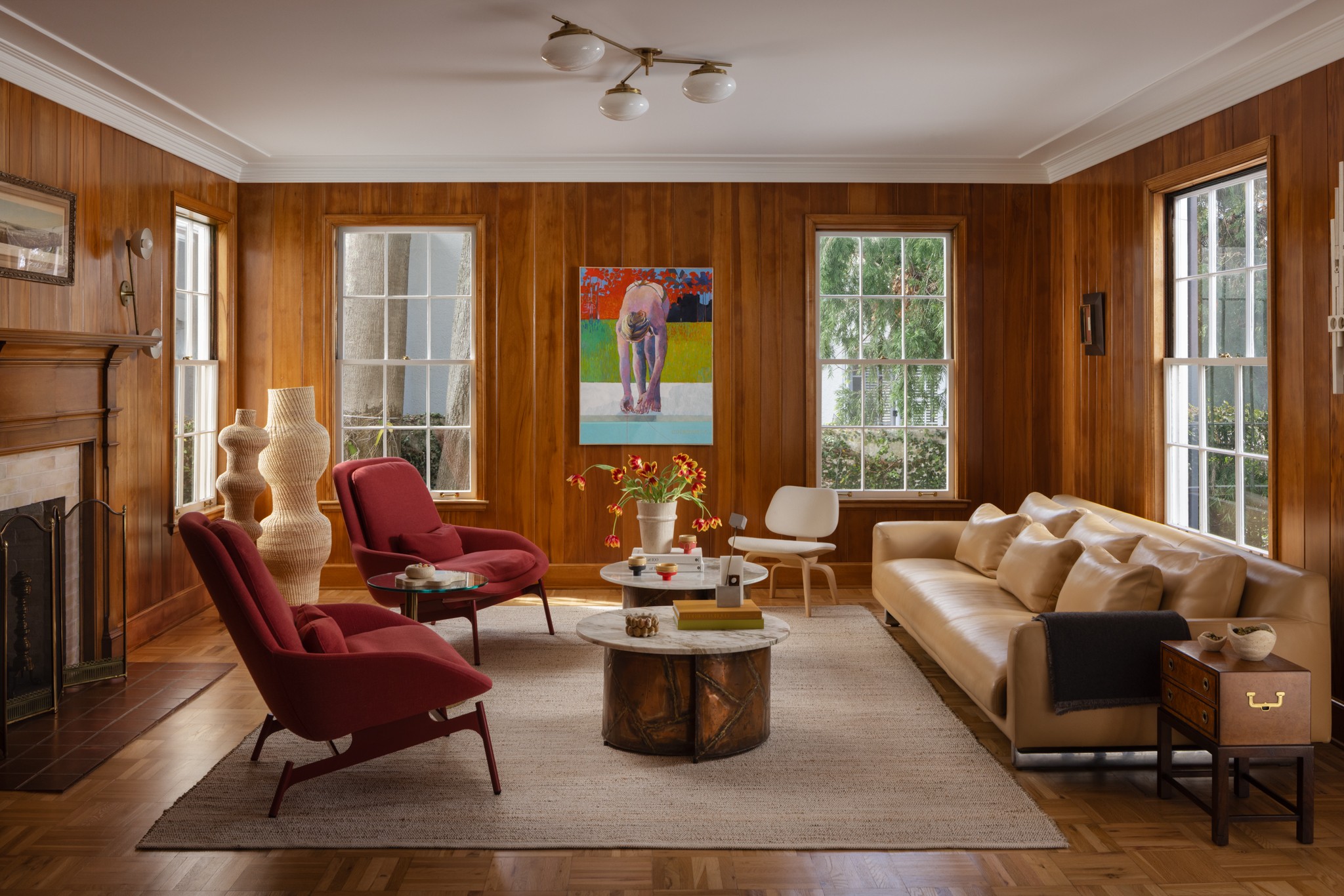 Living room with wood-paneled walls, three windows, a beige sofa, two red armchairs, a white chair, a round wooden coffee table, and artwork on the wall.
