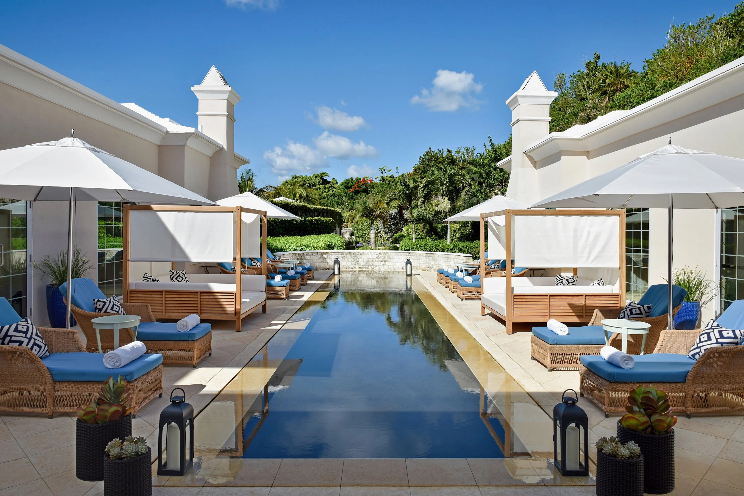 Swimming pool with lounge chairs and umbrellas, white buildings, greenery, blue sky, and clouds in the background.
