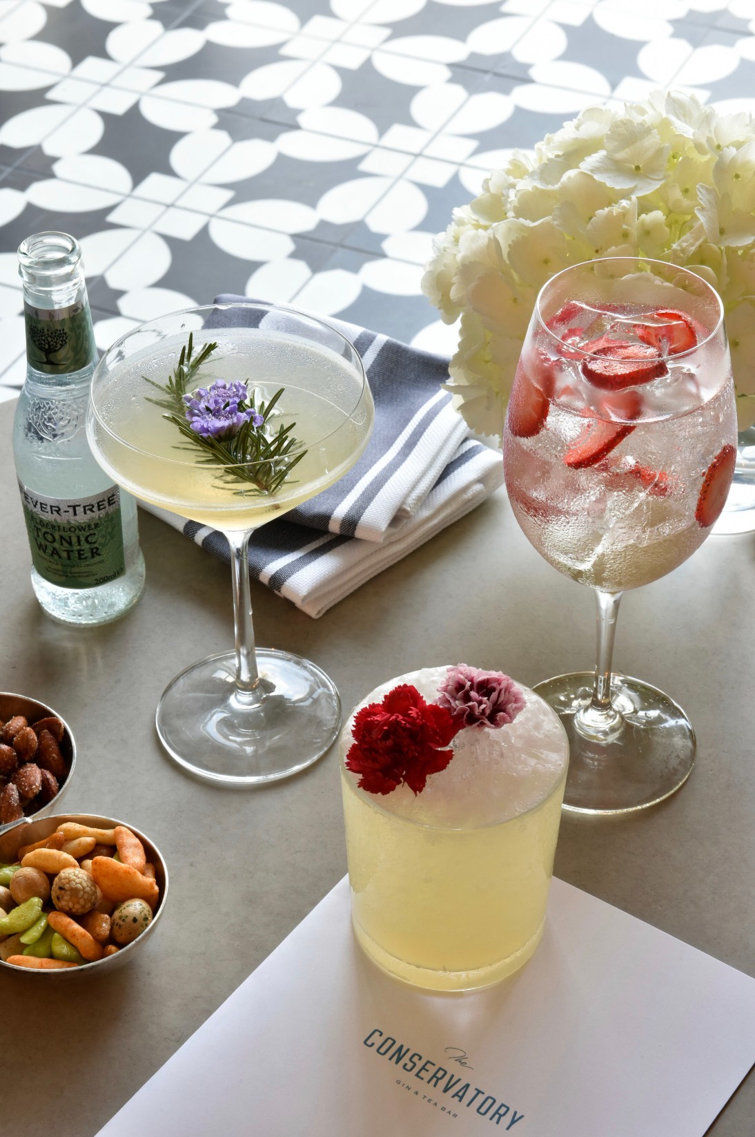 Two cocktails with flowers and ice, a bottle, a bowl of nuts, and a white hydrangea on a patterned table.