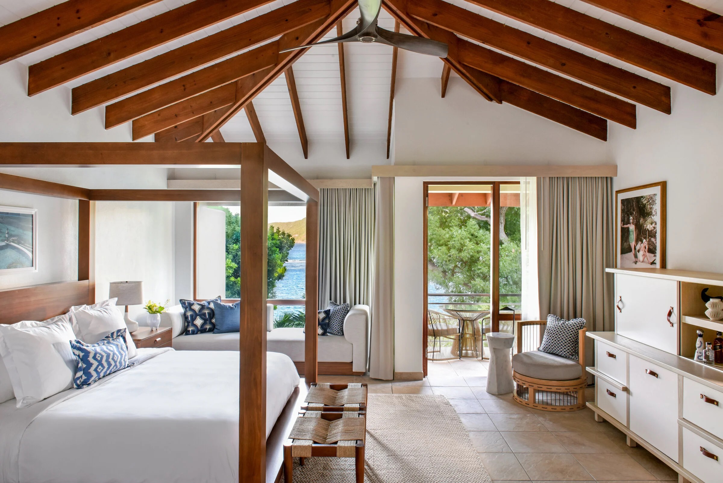 Bright bedroom with wooden ceiling beams, large windows, and a balcony view, featuring a bed, chairs, and white furniture.