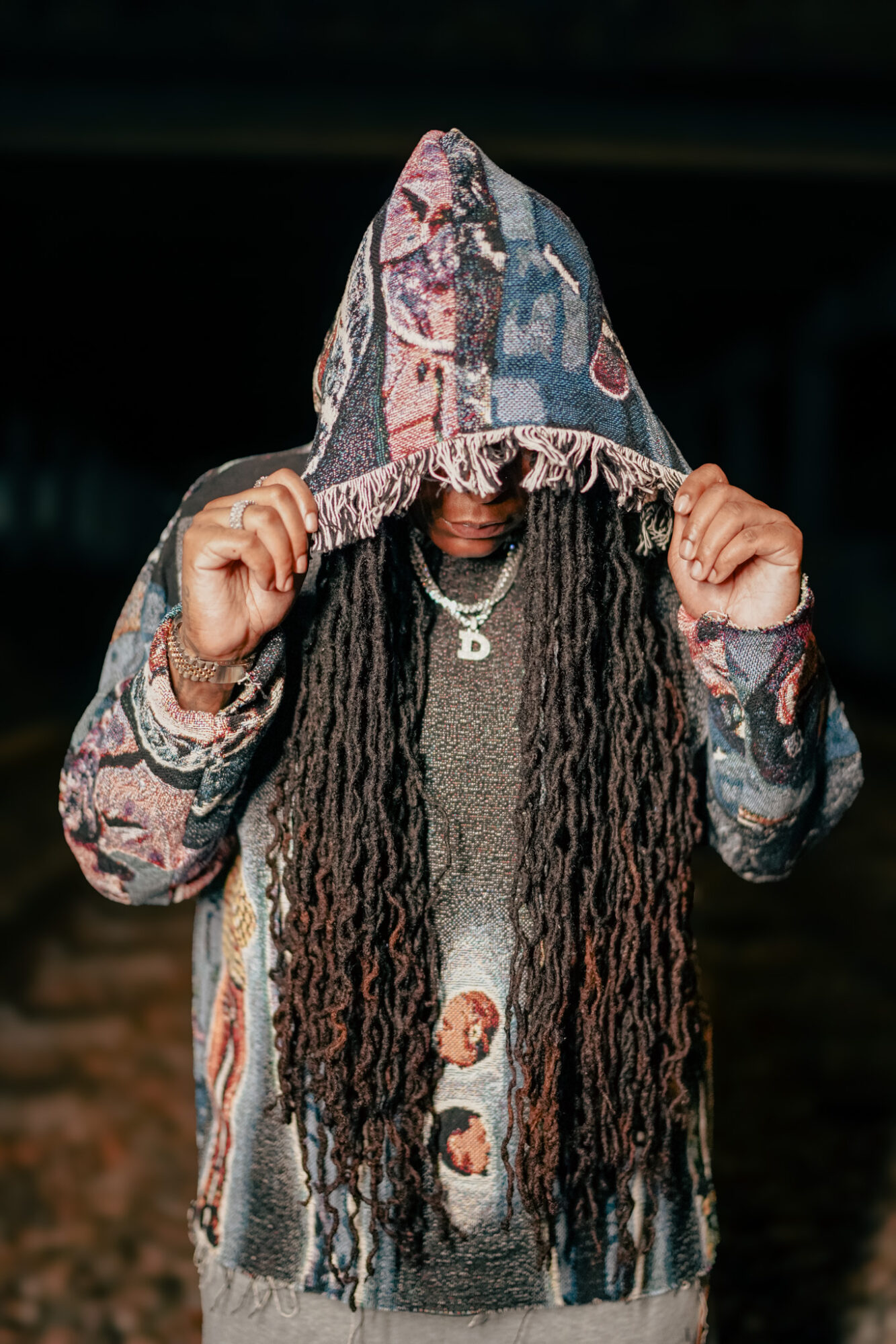 Person with long dreadlocks wearing a patterned hoodie, holding the hood with both hands, face partially obscured.