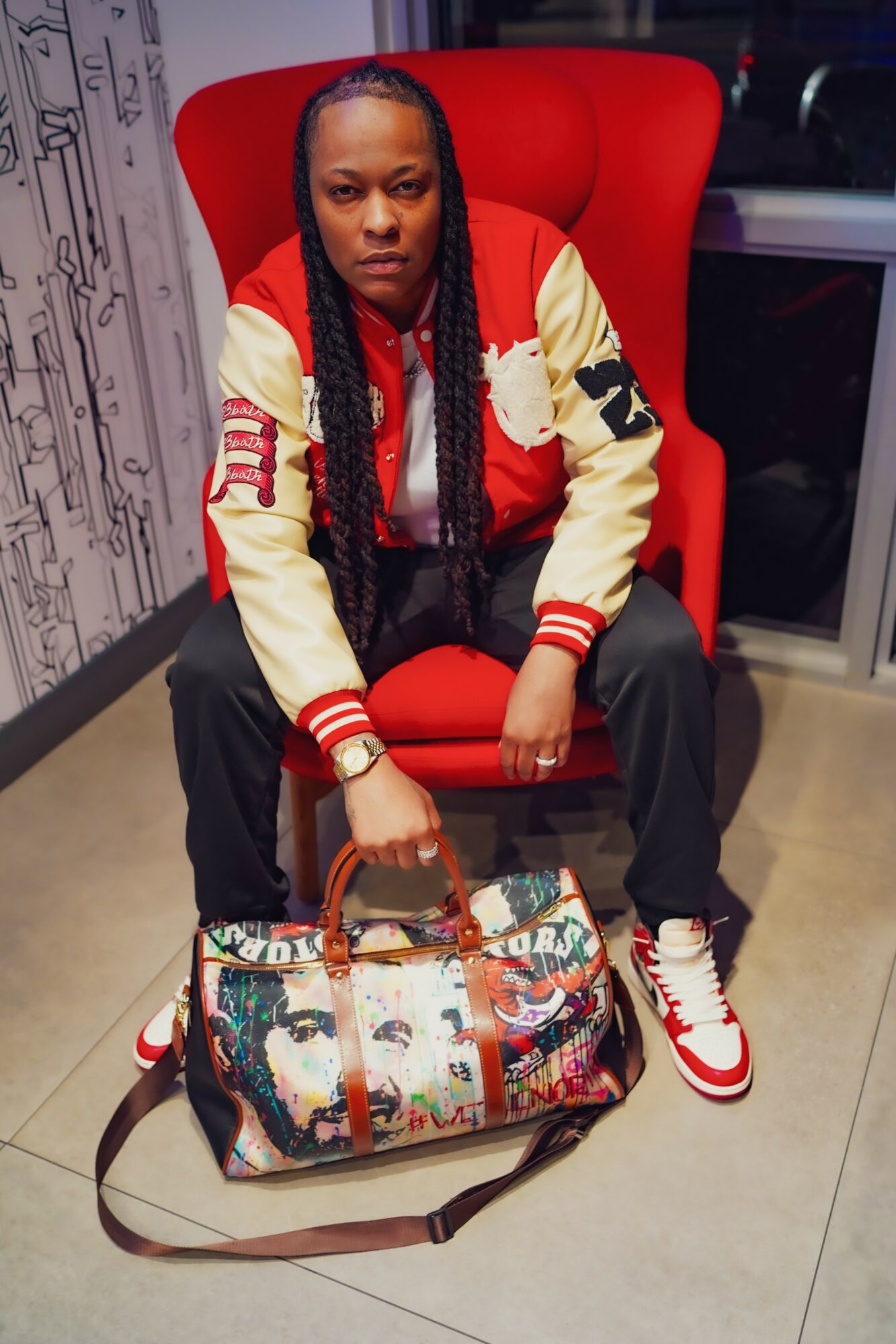 Person with long braided hair sitting in a red chair, holding a colorful bag, wearing a red and cream jacket, black pants, and red sneakers.