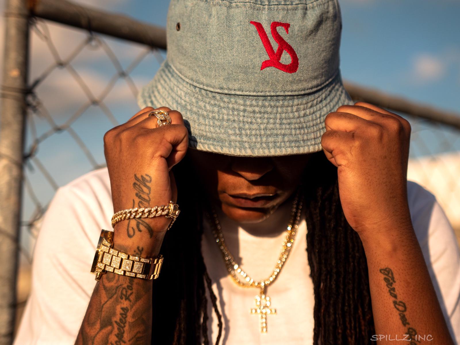 Person wearing a gray bucket hat with red logo, holding the hat with both hands, outdoors with a chain-link fence background.