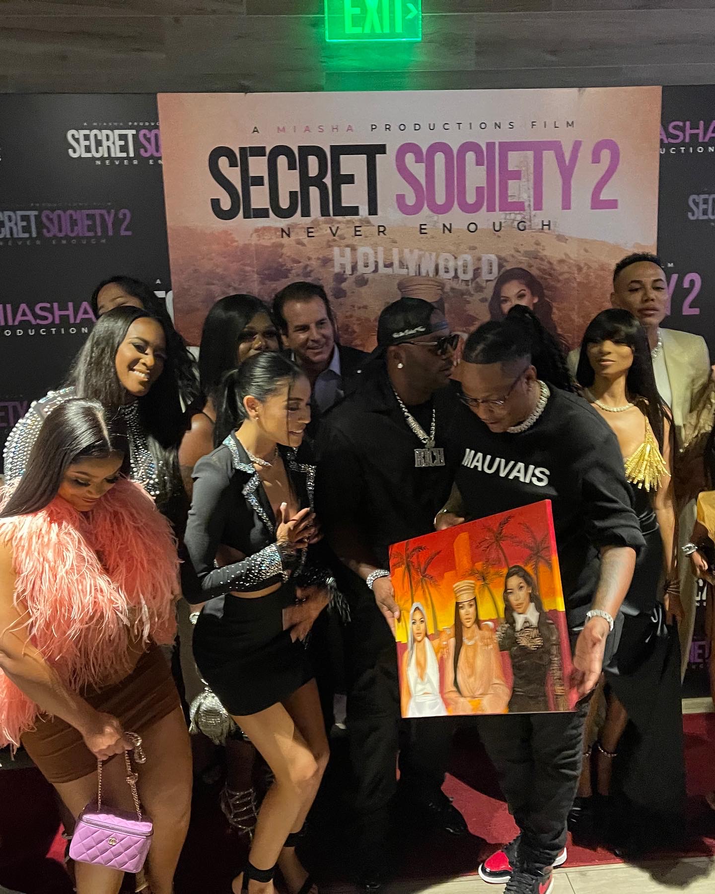 Group of people at a promotional event holding a colorful painting, with a backdrop for 'Secret Society 2' movie.