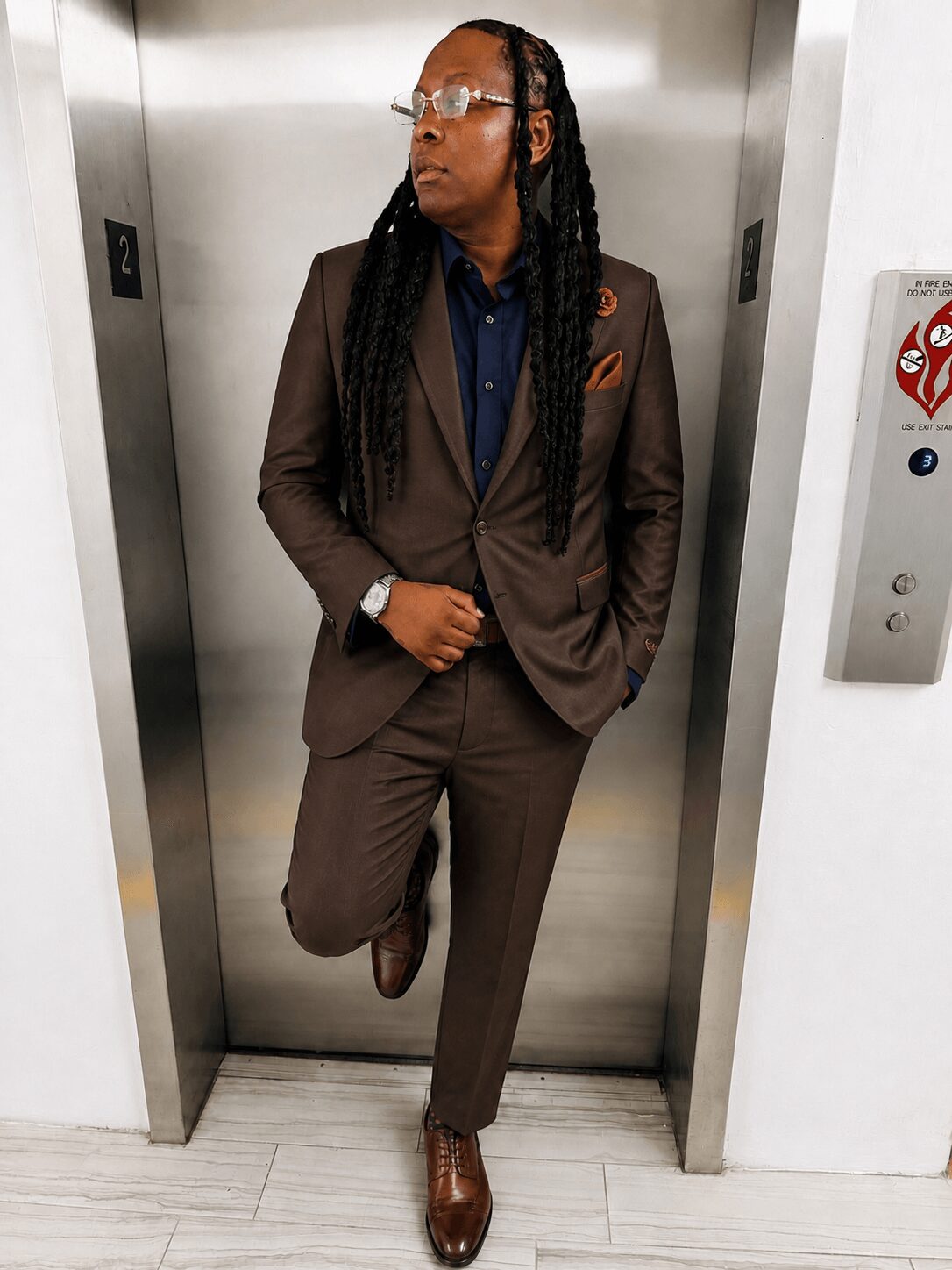 Person in a brown suit with glasses and long braids standing in an elevator, leaning against the wall.