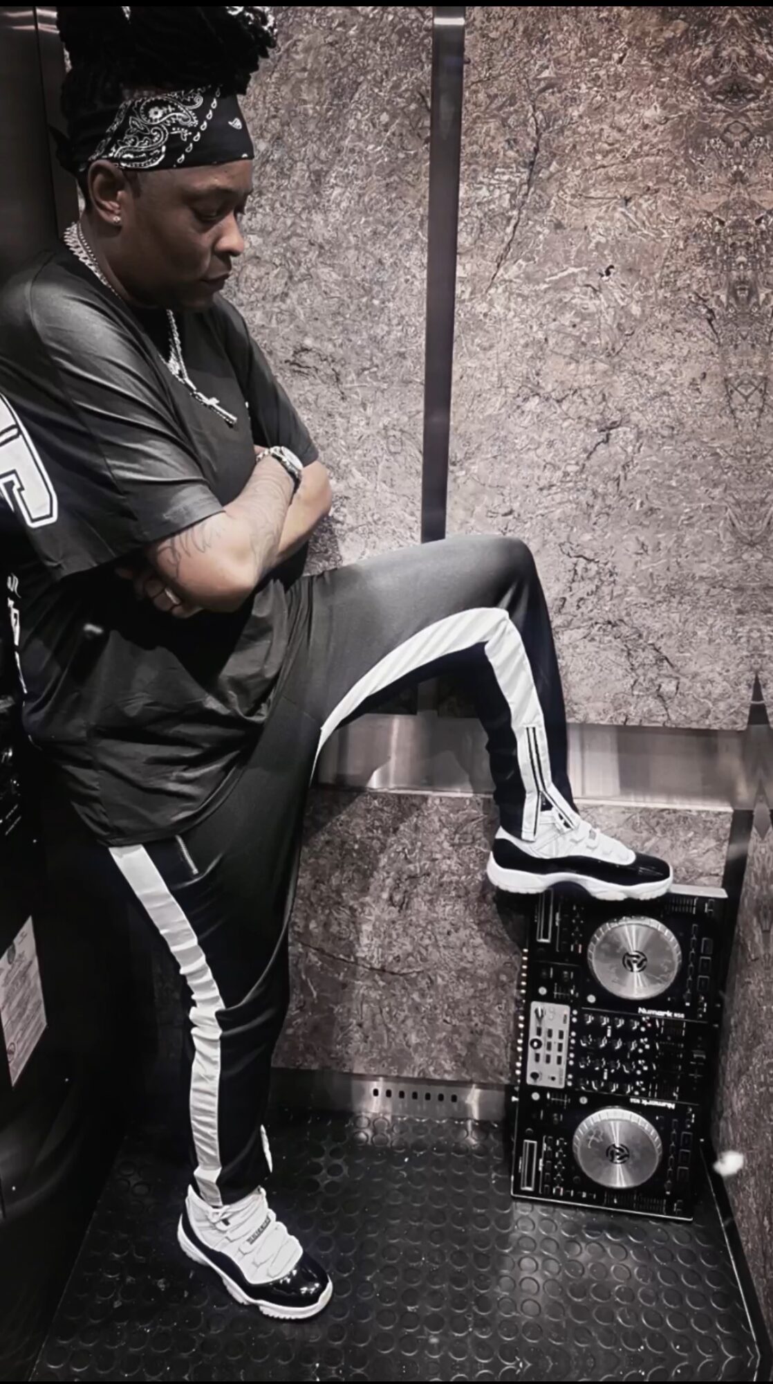 Person with arms crossed, wearing black sportswear, standing with one foot on a speaker, in front of a textured wall.