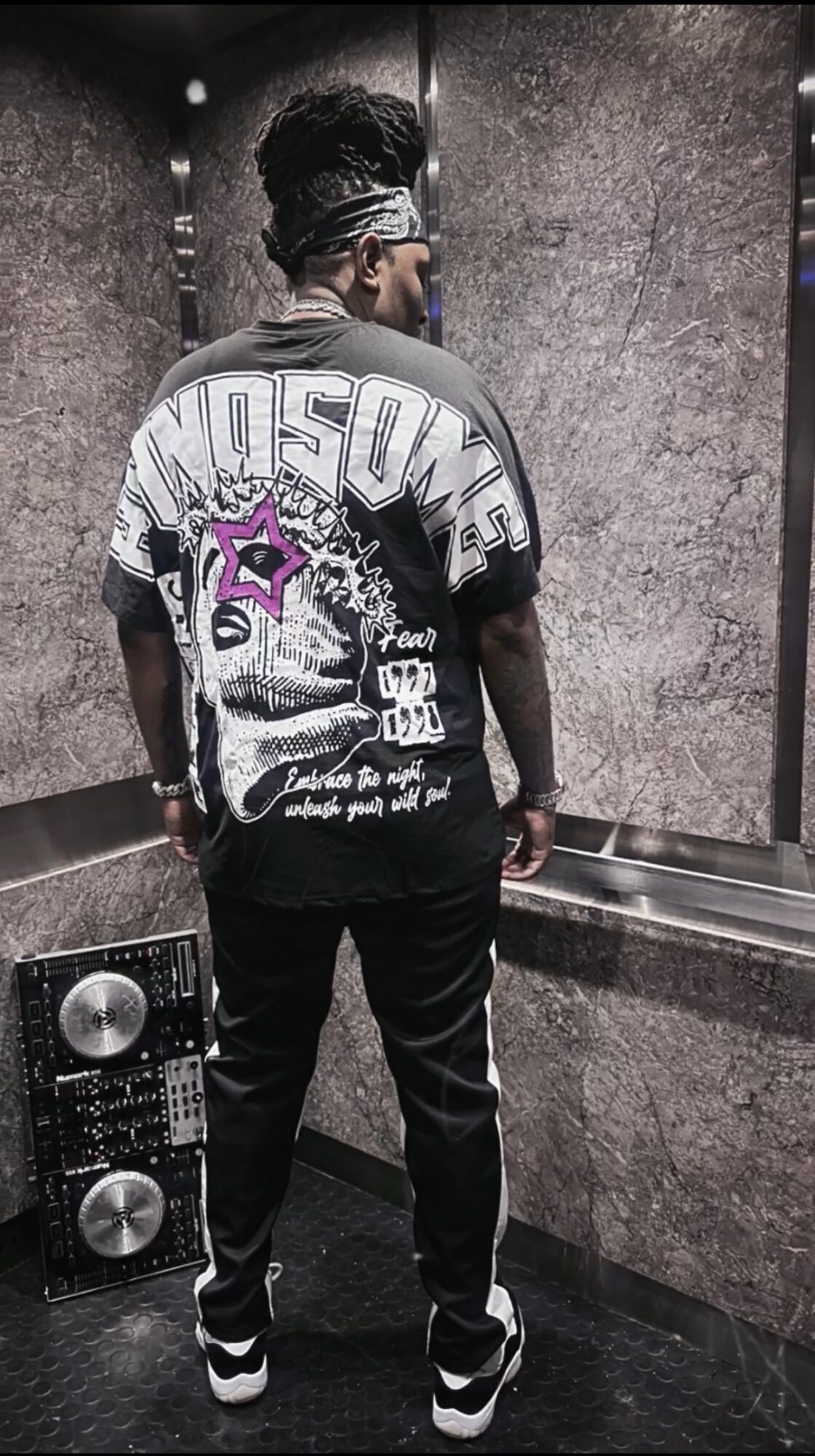 Person standing with back to camera in an elevator, wearing a graphic T-shirt, black pants, and sneakers.