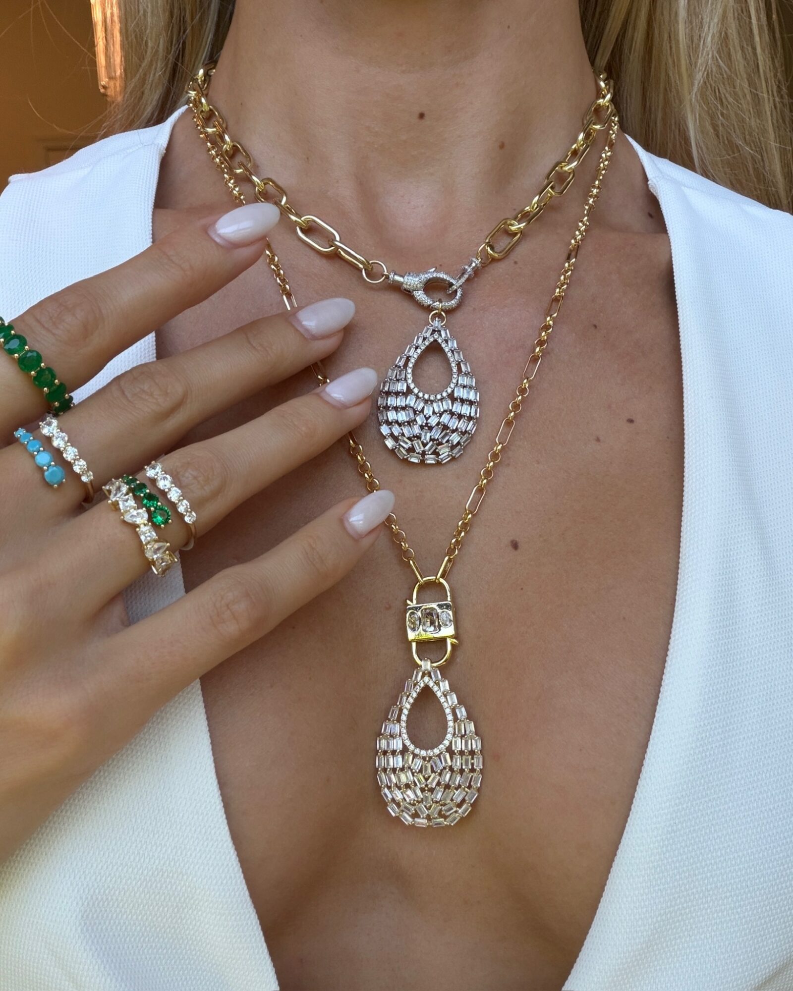 Close-up of a woman wearing layered gold and diamond necklaces, with her hand showing multiple rings and a white top.