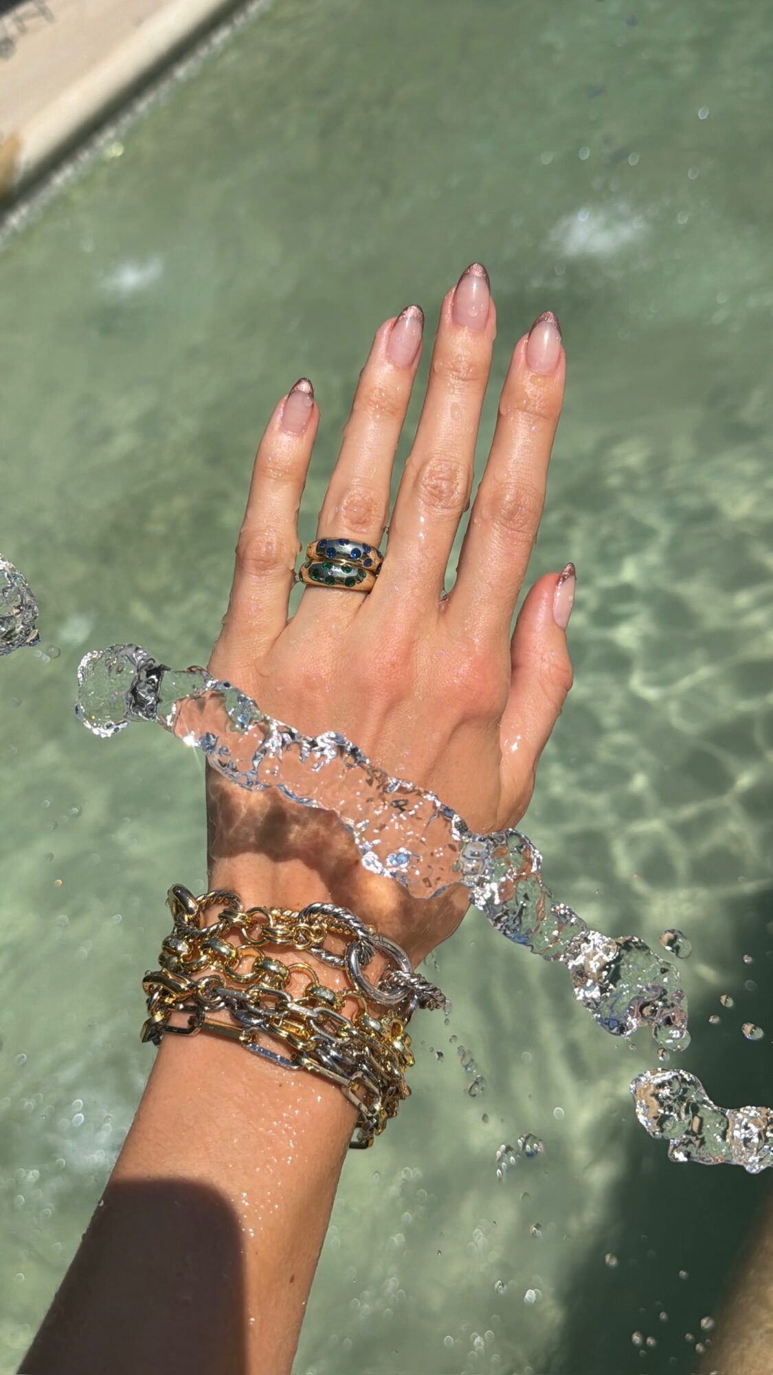 Hand with jewelry submerged in water, with water splashing around fingers, greenish background, multiple bracelets, and a ring.