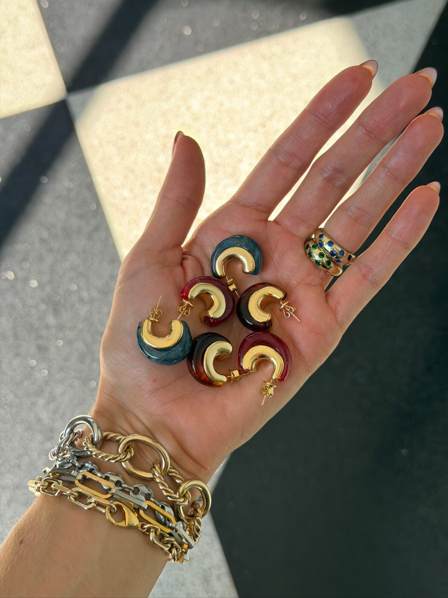 Hand with multiple gold and black earrings, wearing gold bracelets, against a background with sunlight and shadows.