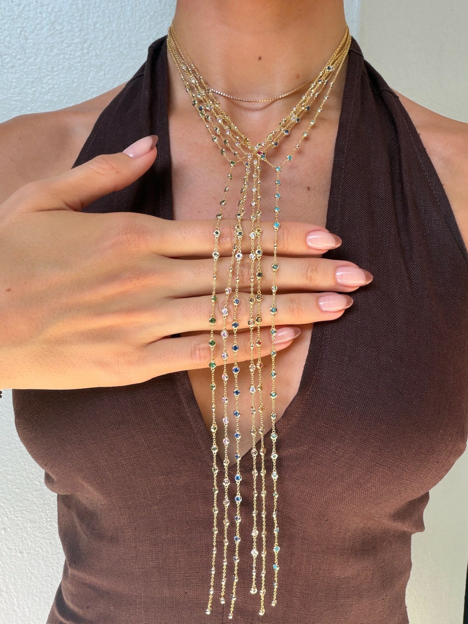 Person wearing layered gold necklaces and a brown top, with hand resting on chest, against a plain background.