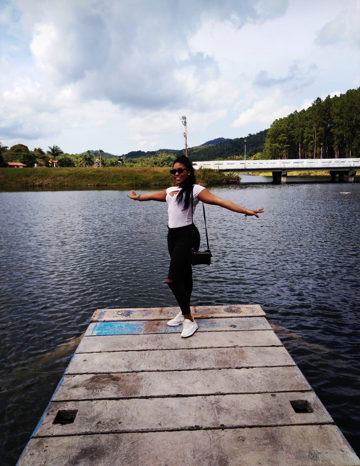Person standing on a wooden dock with arms outstretched, water and bridge in background, cloudy sky, wearing sunglasses, white shirt, black pants, white shoes, black bag