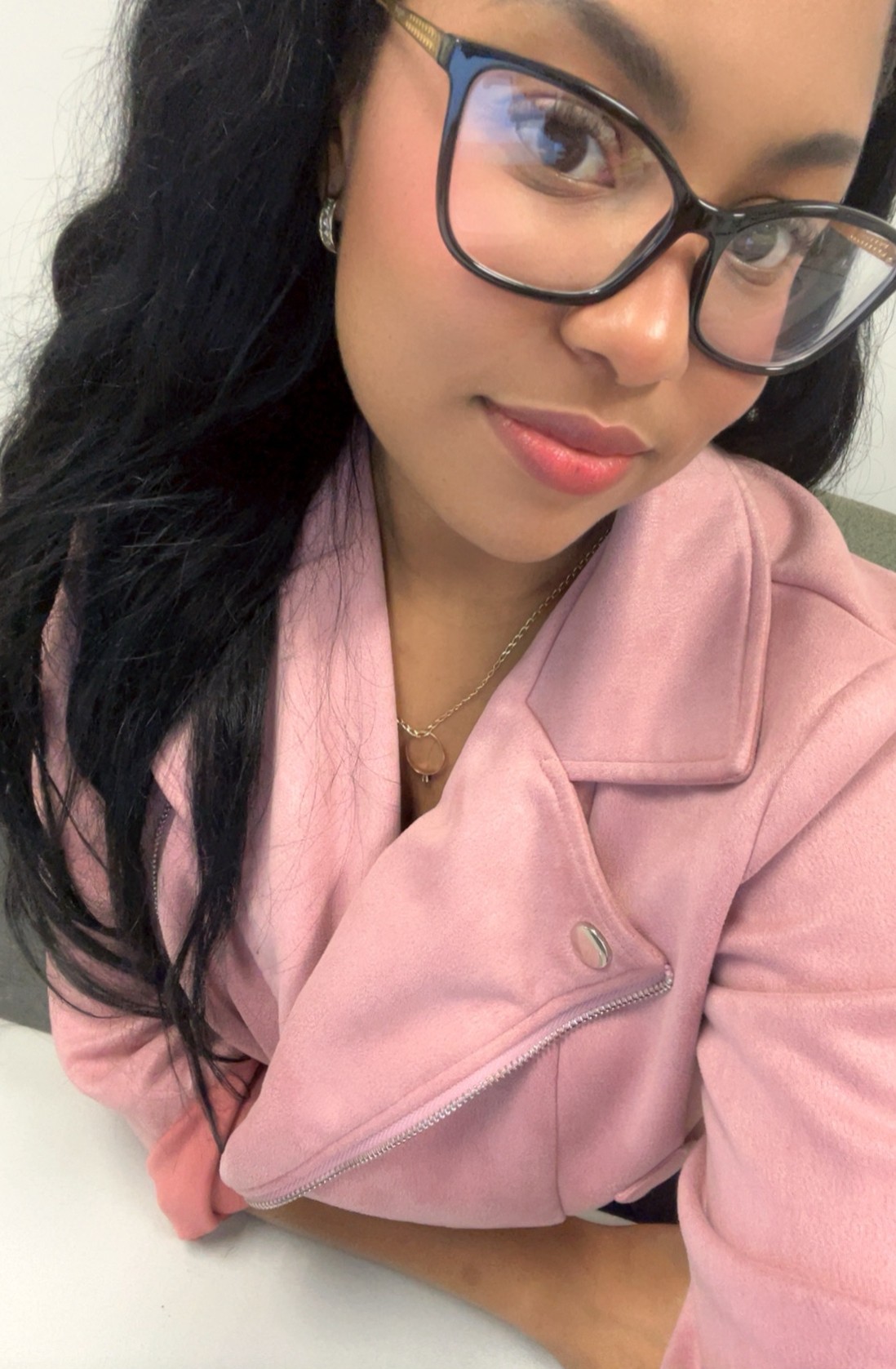 Close-up of a woman with glasses, long dark hair, wearing a pink jacket, looking at the camera.