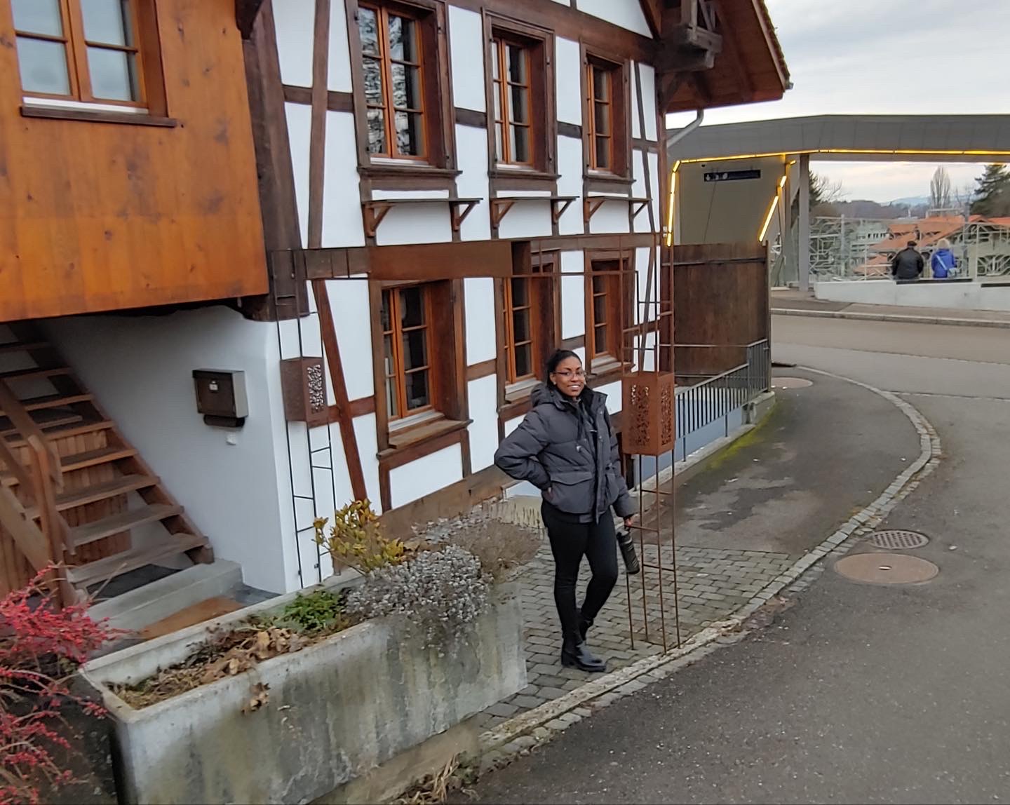 Person standing on sidewalk near a traditional wooden and white building, smiling, wearing a dark jacket and glasses.