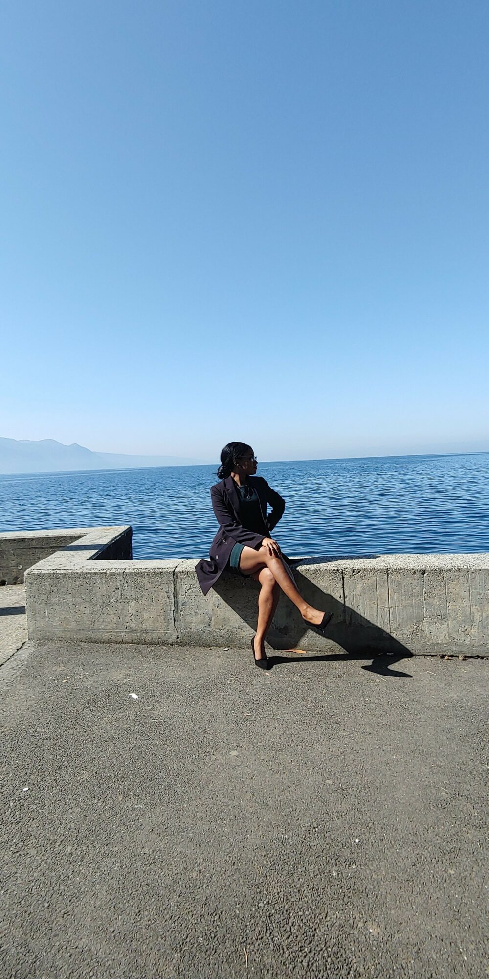 Person sitting on a concrete ledge by the ocean, facing away, with a clear blue sky and distant mountains.