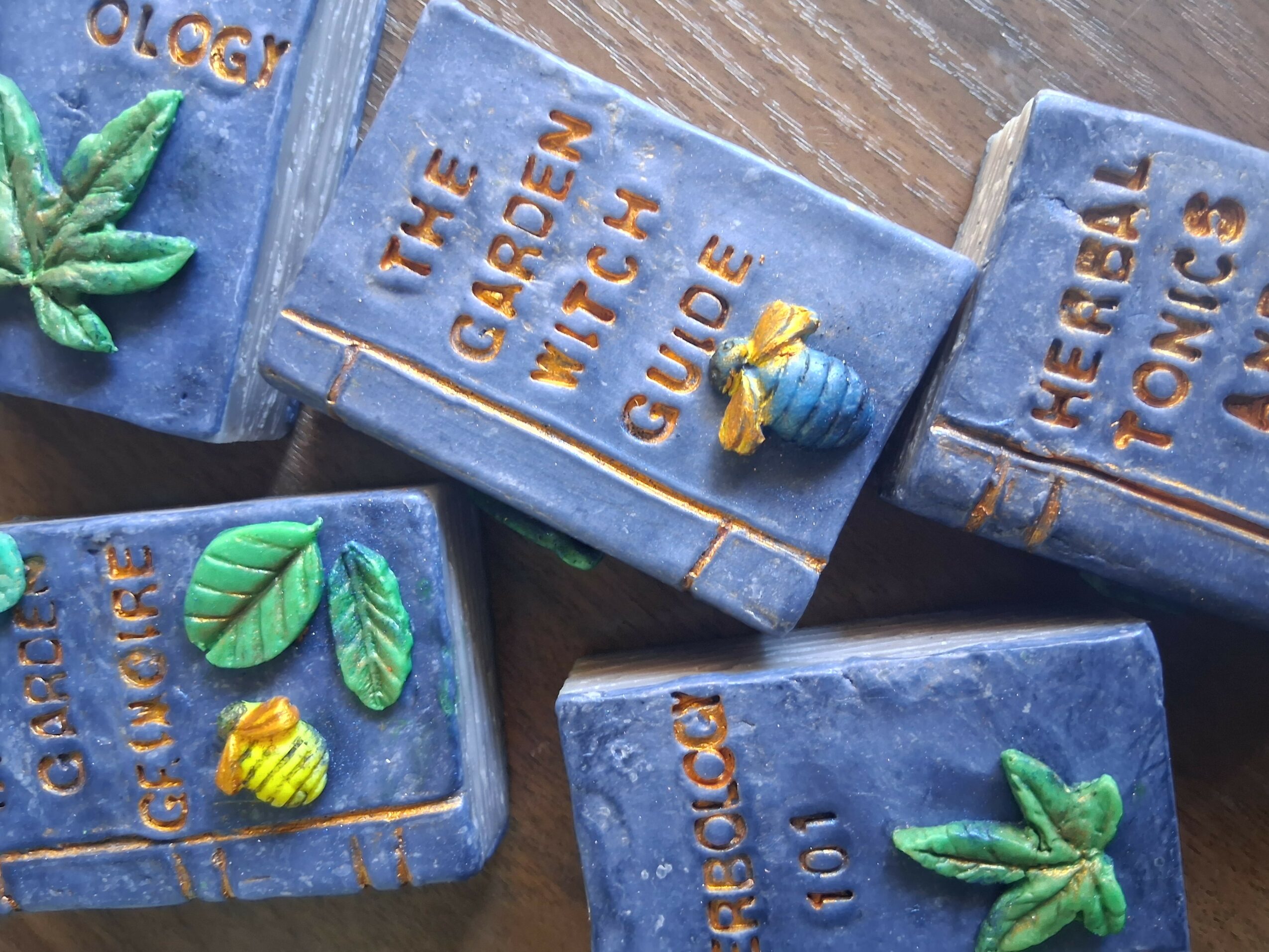 Four blue soap bars with colorful plant and bee decorations, arranged on a wooden surface.