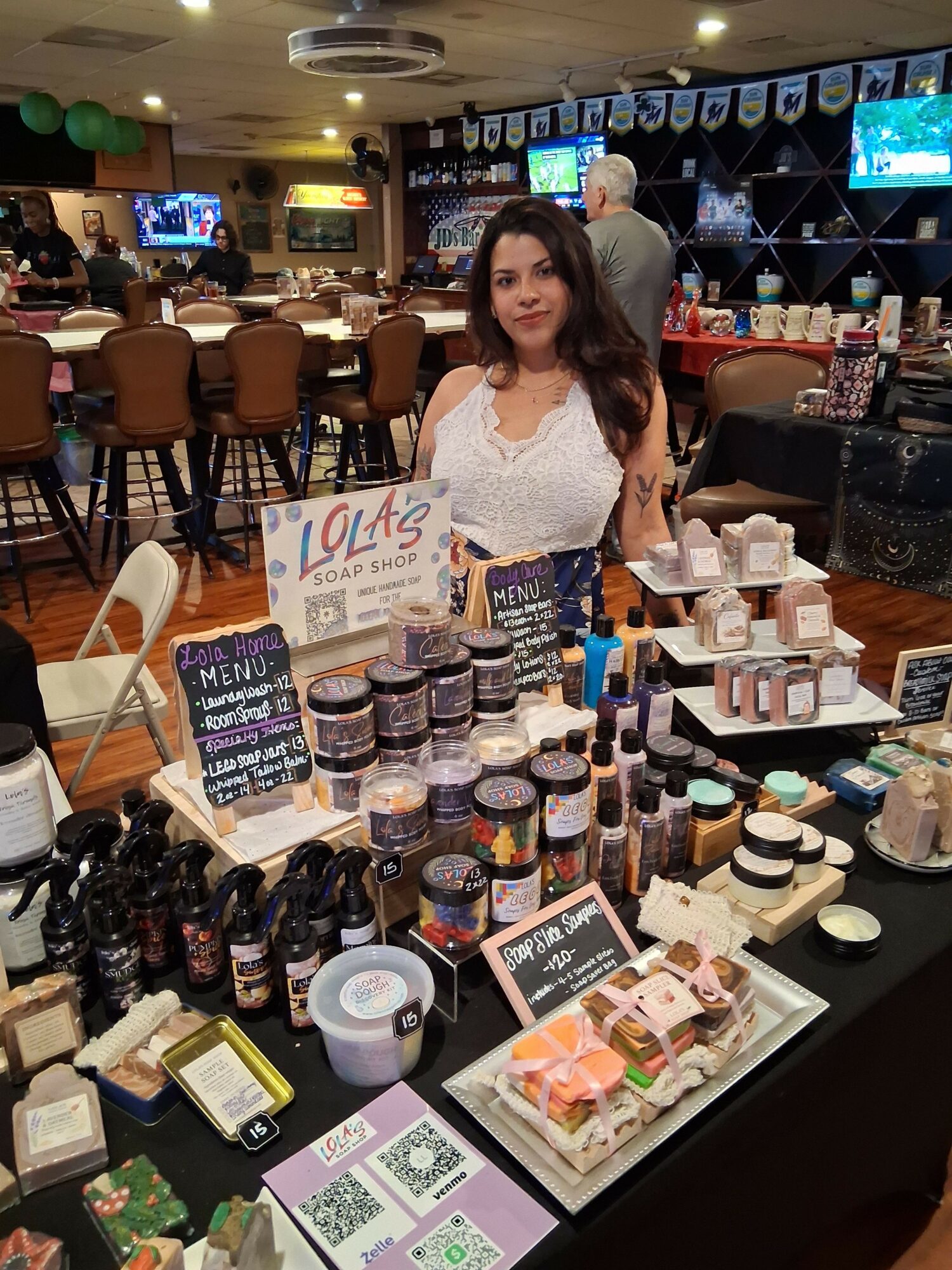 Woman standing behind a table with various beauty products, signs, and promotional materials in a store or market setting.