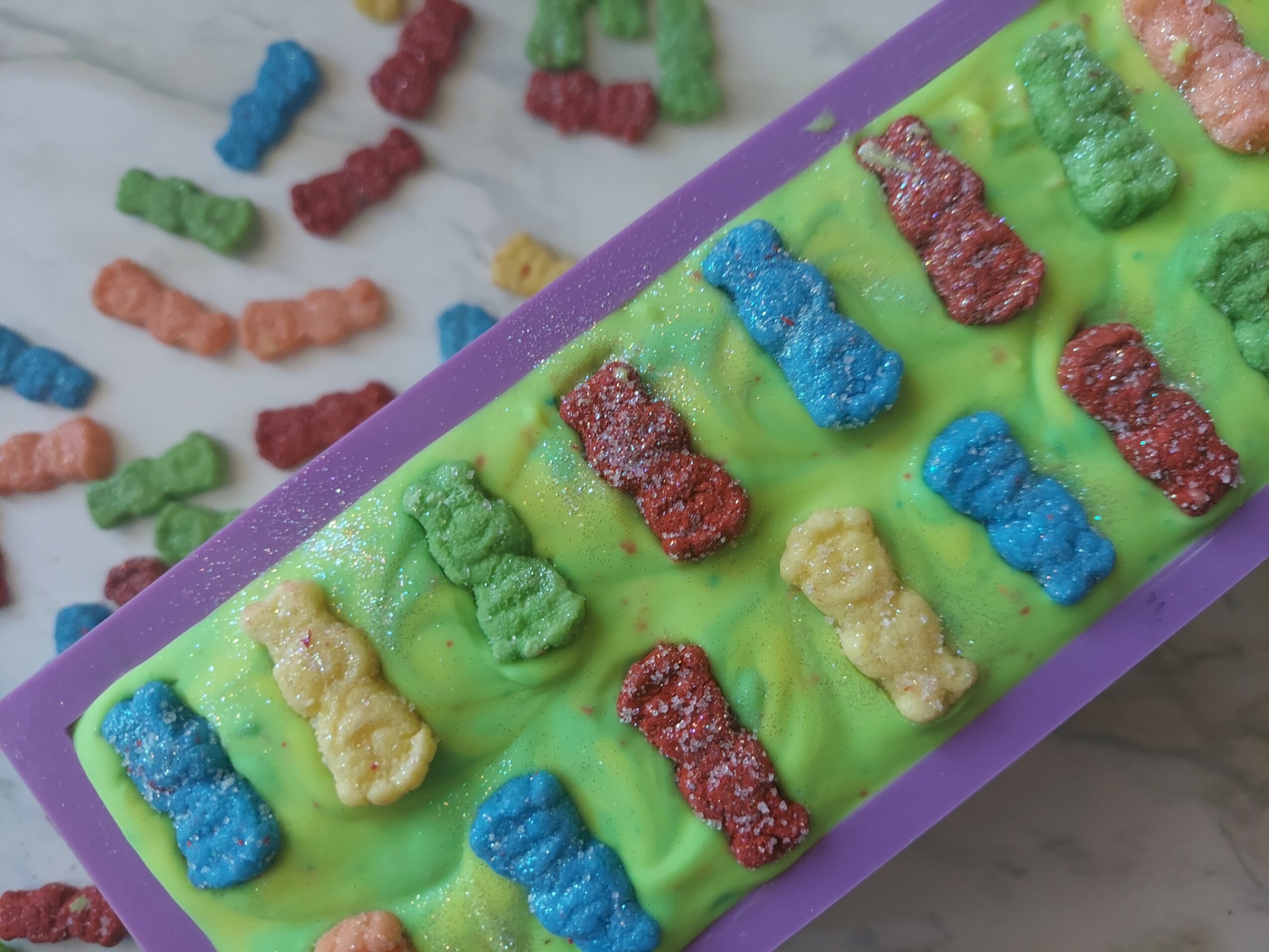Colorful gummy bears arranged in a row on a green surface with scattered gummy bears nearby, purple border visible.