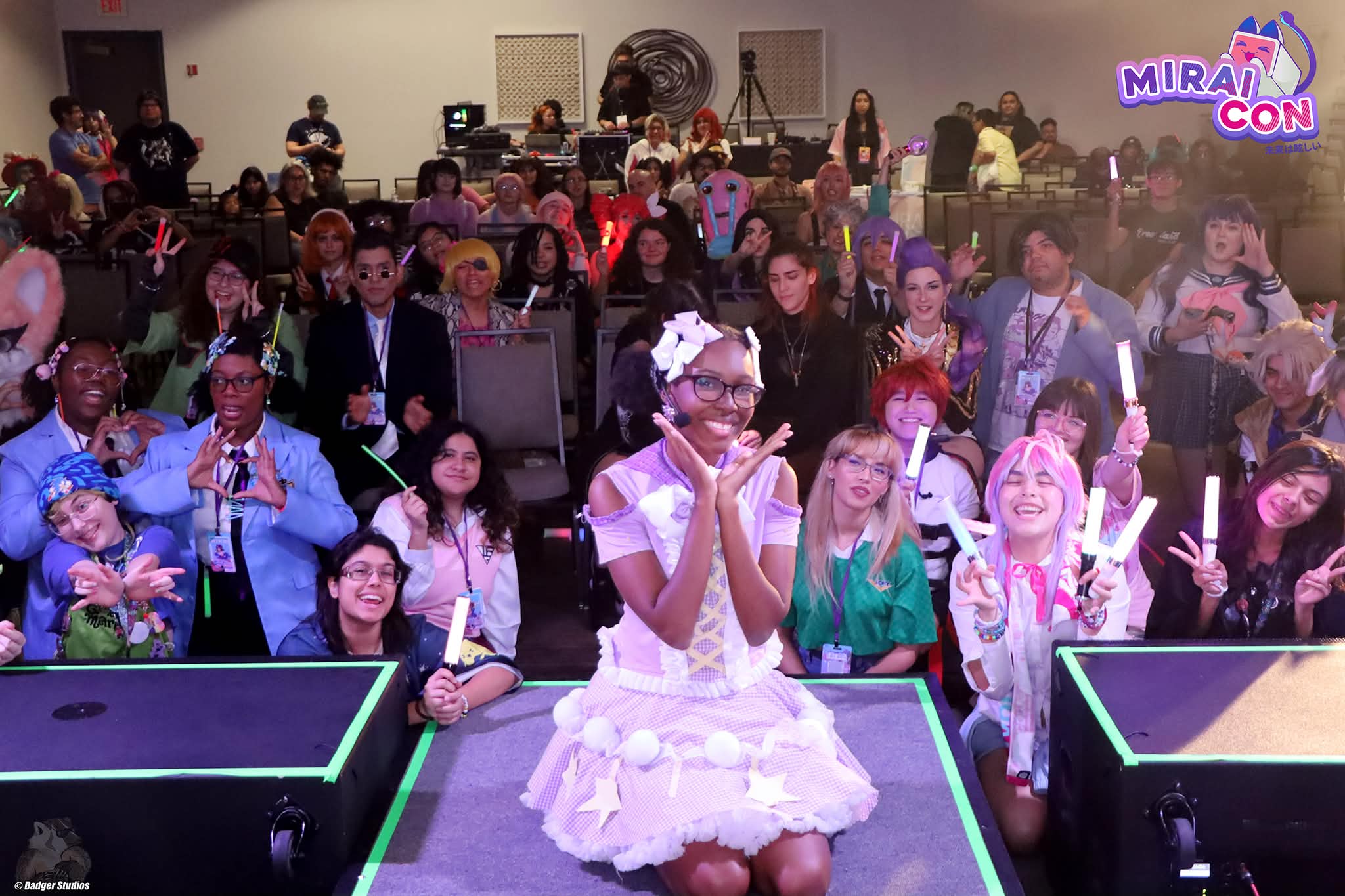 Group of children and adults at a gaming event, some holding glow sticks, with a woman in a white dress in front.