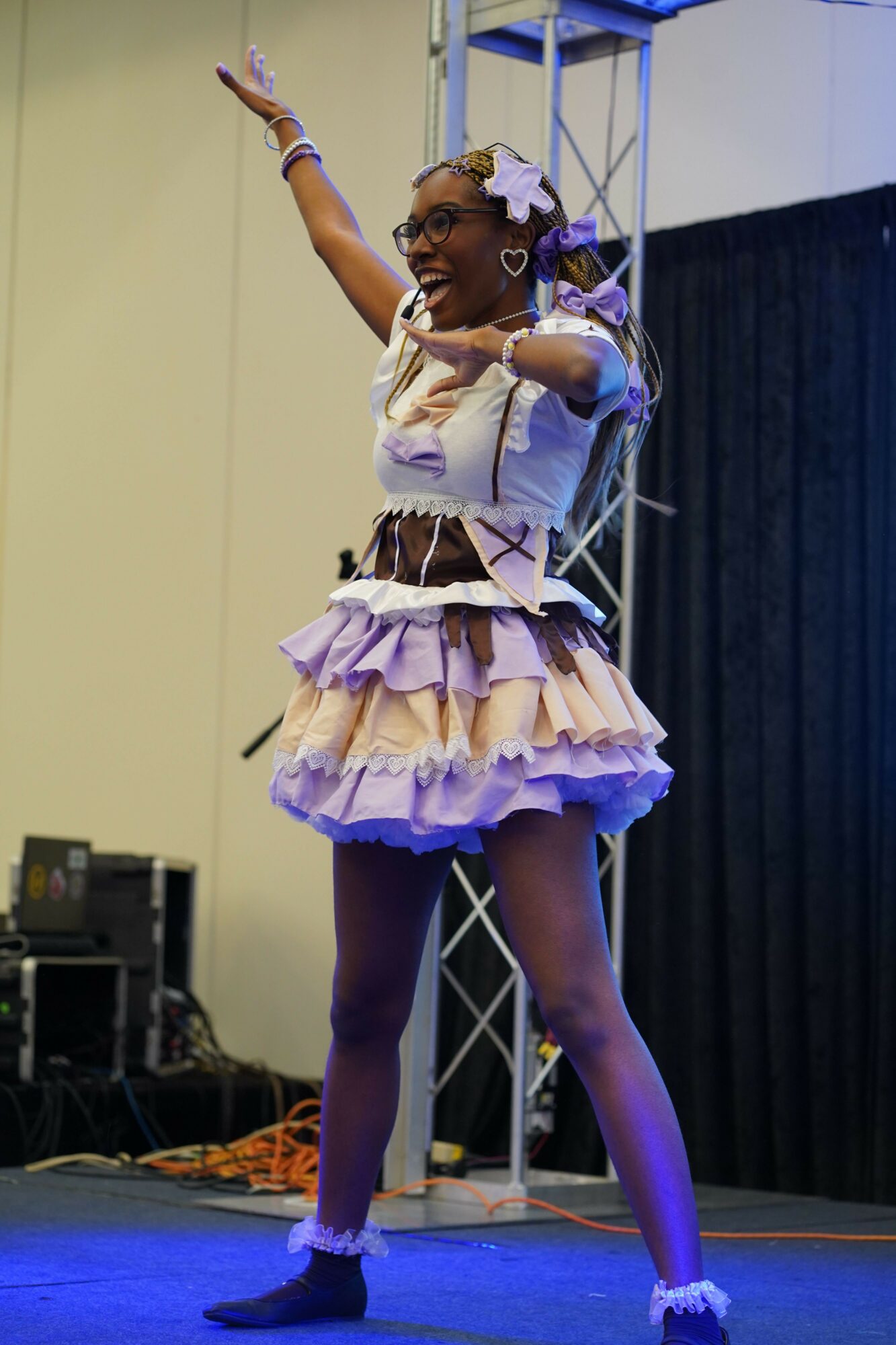 A young girl performs a dance move on stage, wearing a purple and white costume with ruffles and a bow.