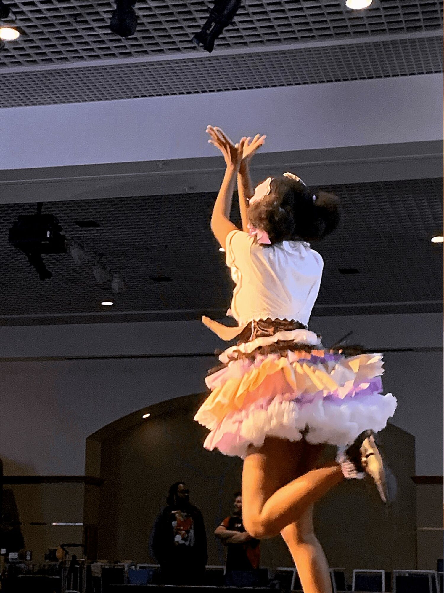 A person dancing on stage wearing a colorful tutu and white top, with arms raised and one leg lifted.