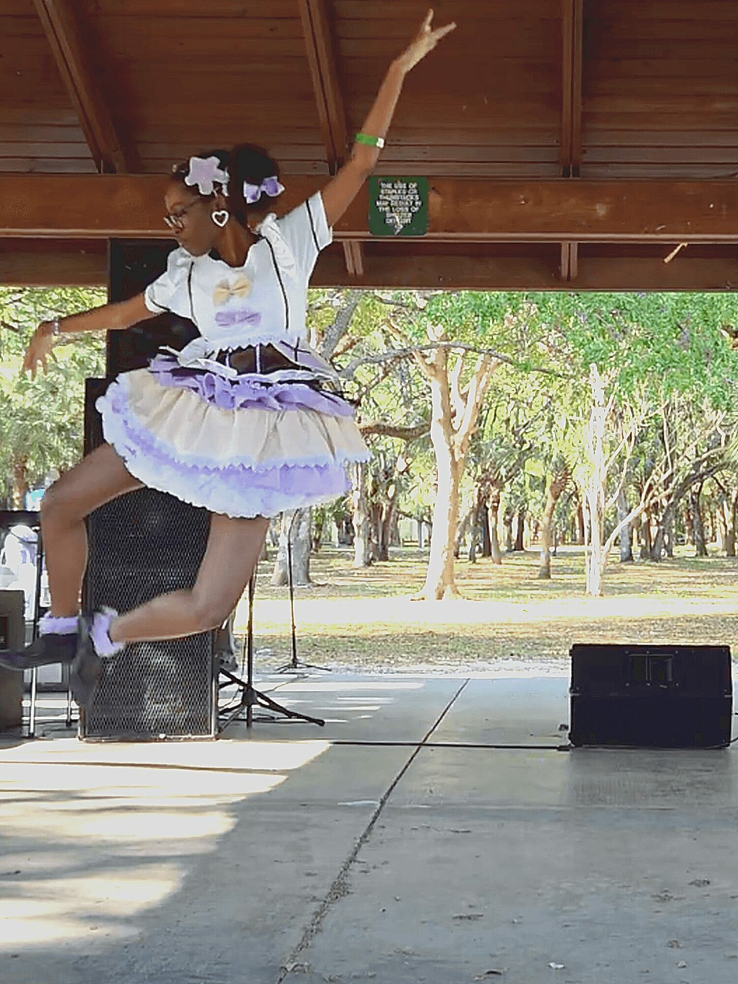 Young girl in a colorful dress dancing on stage outdoors under a wooden roof, with trees in the background.