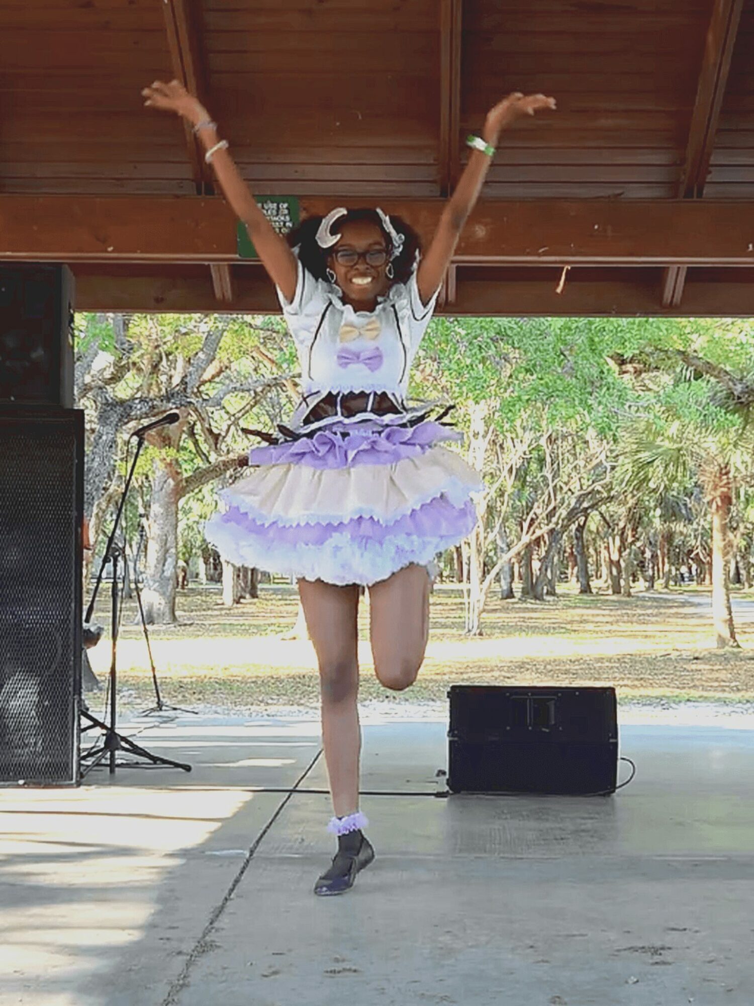 Young girl in a purple tutu and white top jumping with arms raised on stage outdoors, trees in background.