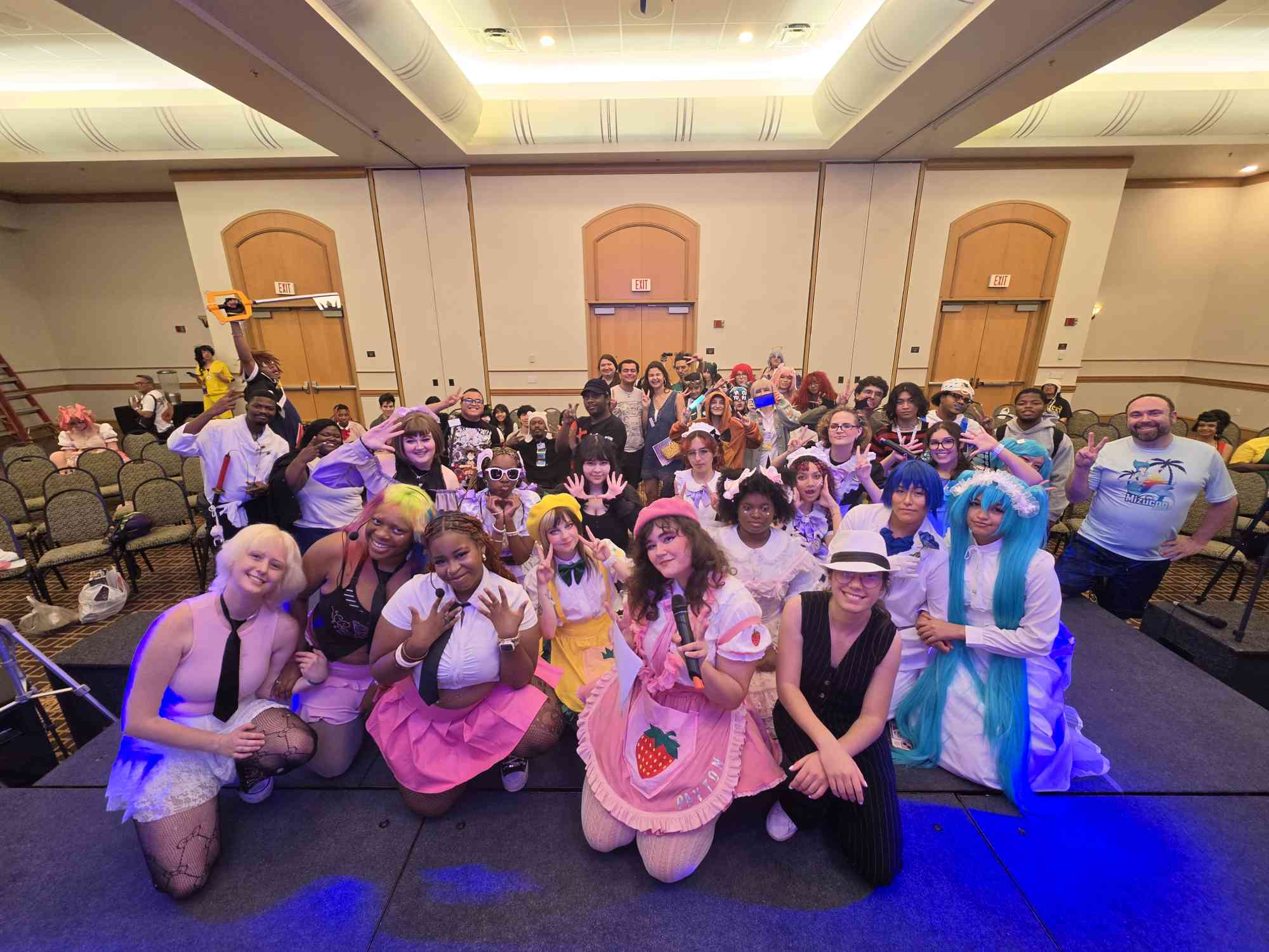 Group of people in colorful costumes posing in a large room with chairs and doors, some kneeling and some standing, smiling at the camera.