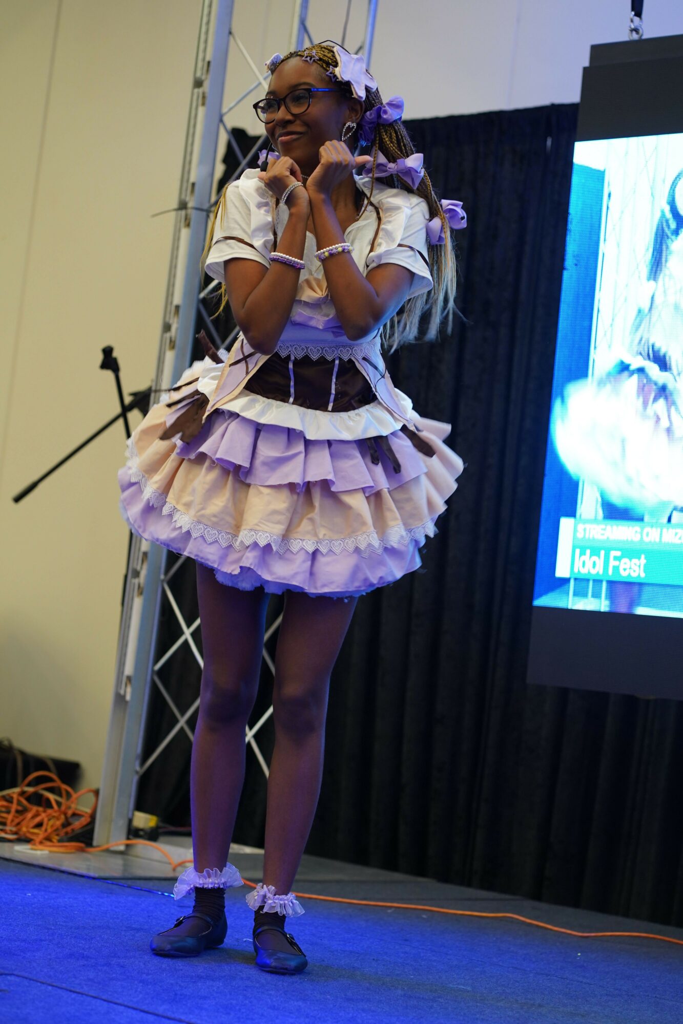Person performing on stage in a ruffled dress, holding a microphone, with a large screen behind them, stage setup visible.