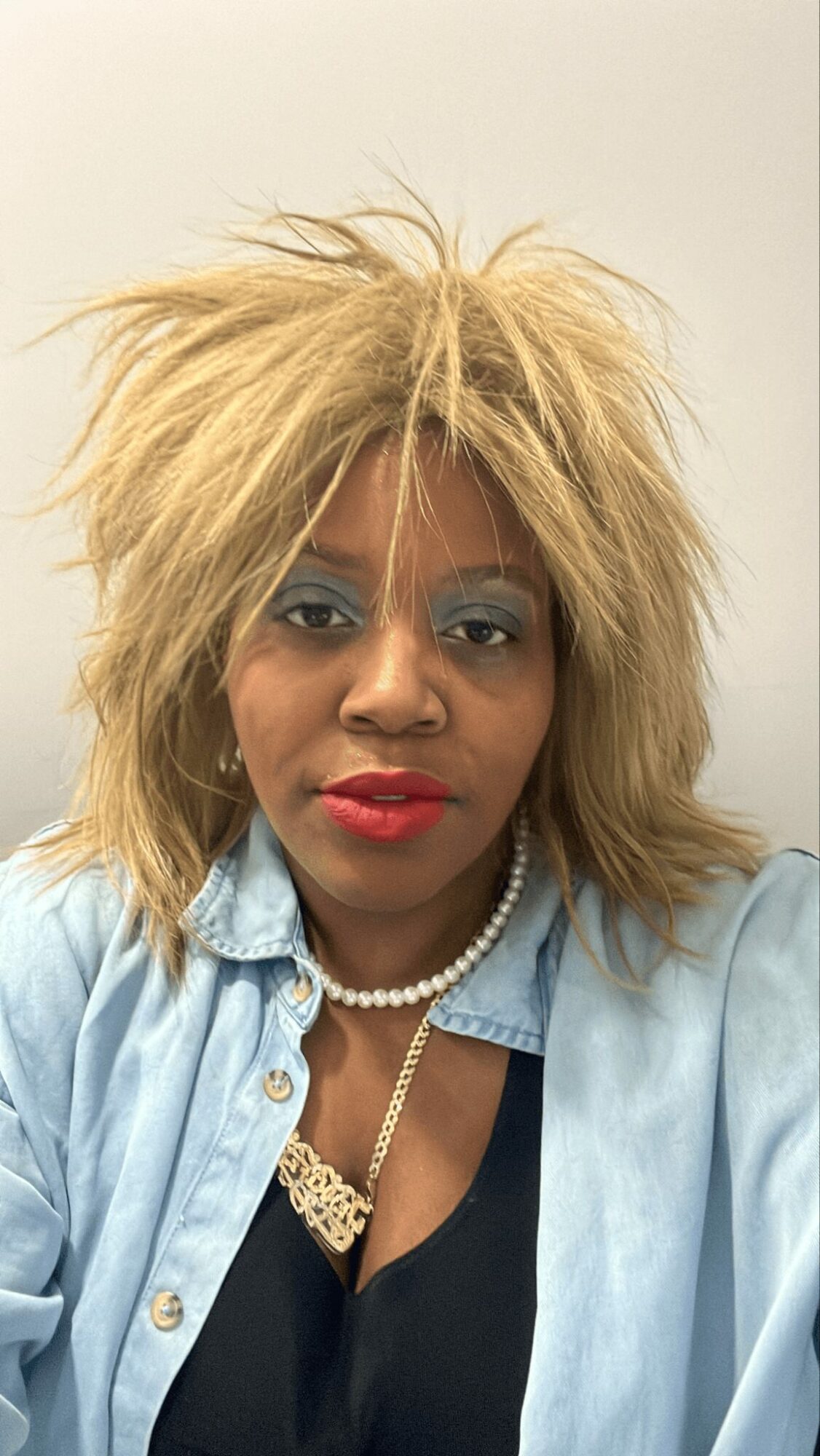 Woman with blonde, messy hair, wearing a denim shirt, pearl necklace, and red lipstick, looking at the camera.