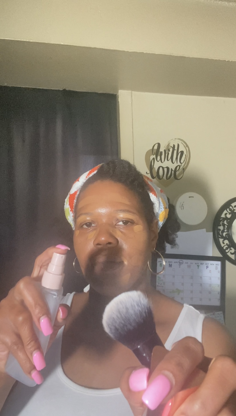 Woman with headband holding makeup brush and spray bottle, pink nails, in room with wall decor and clock