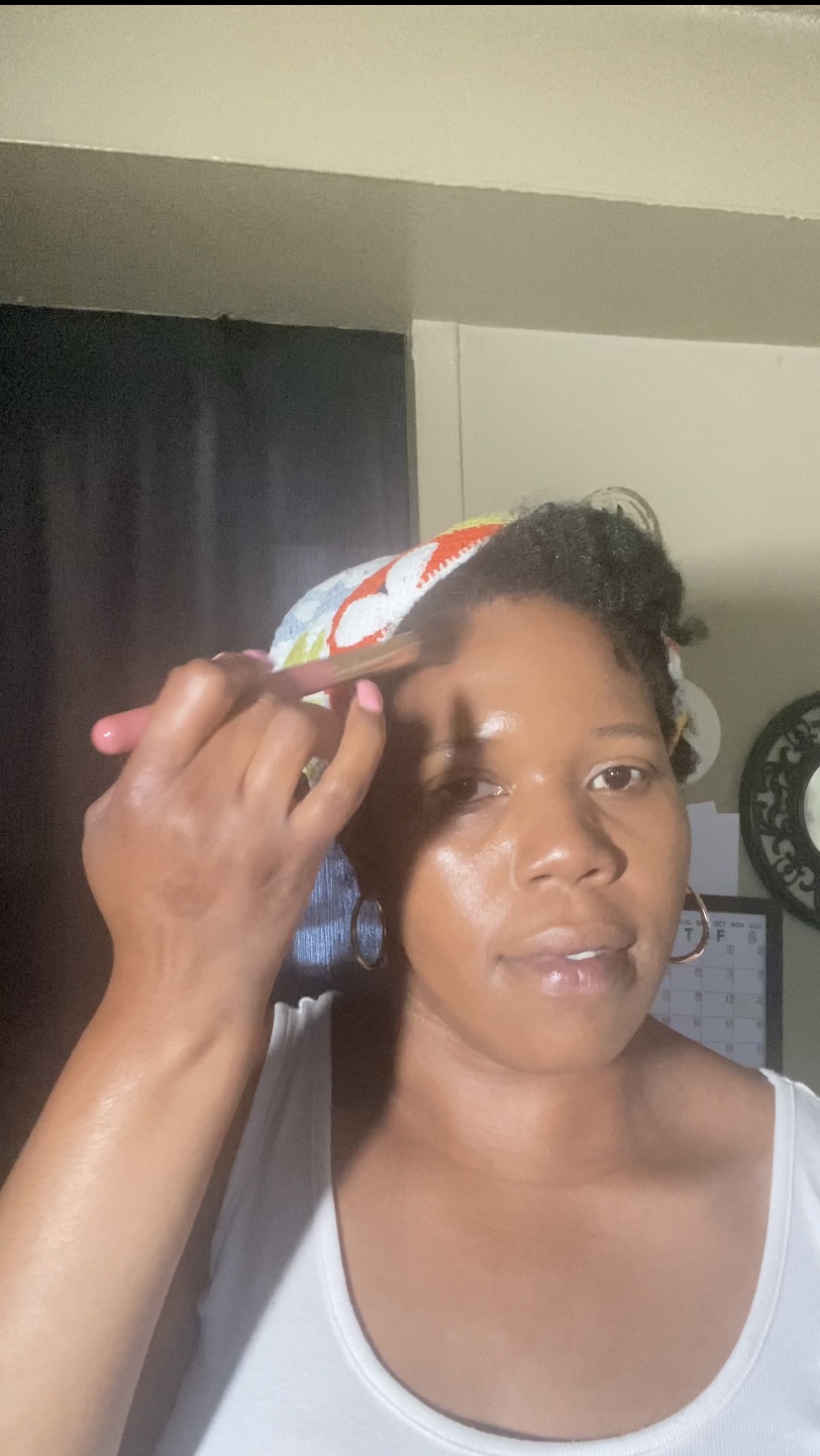 Woman applying makeup with a brush, wearing a colorful headscarf and white tank top, in a room with dark curtains and a clock.