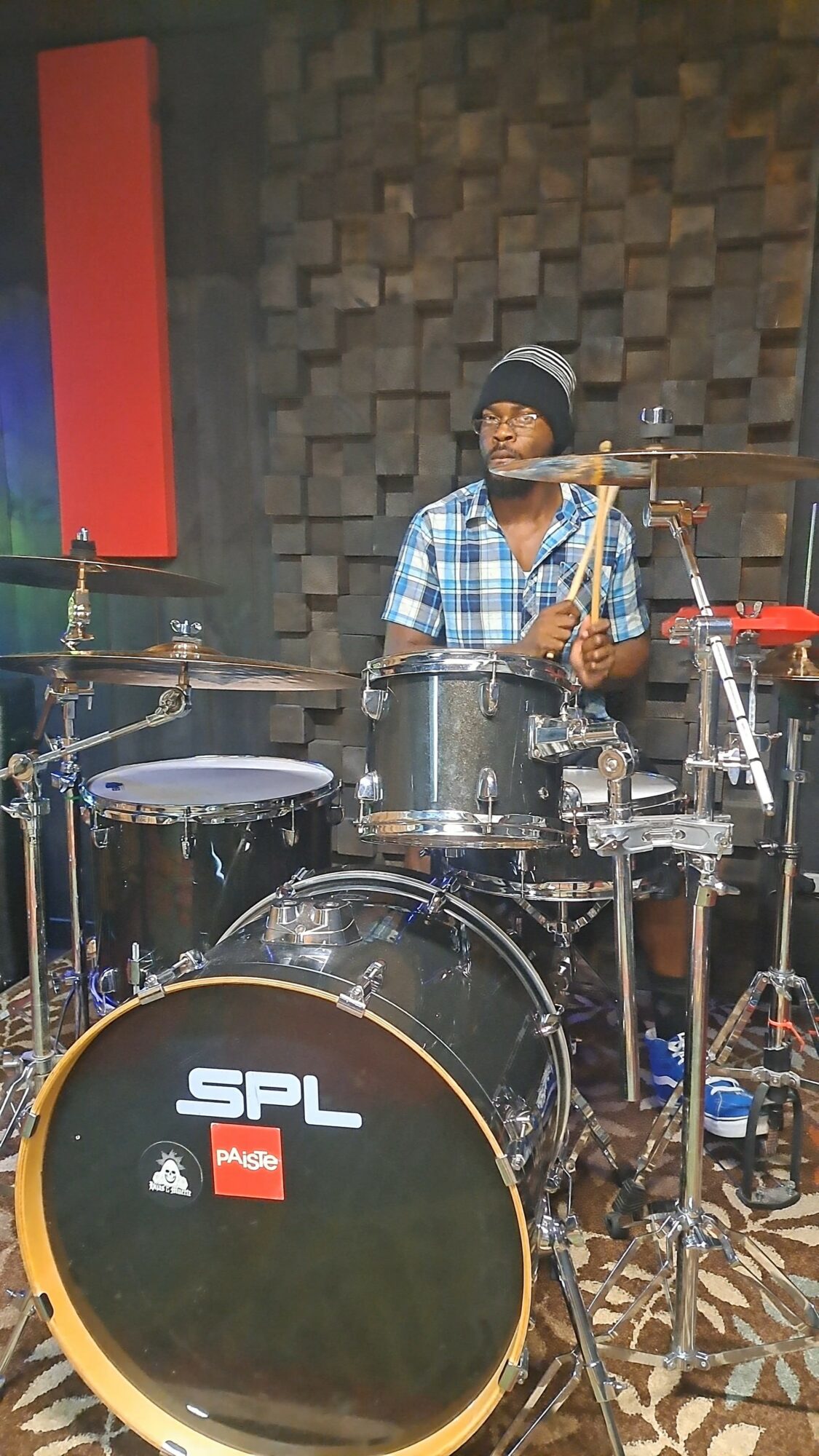 Person playing drums in a music studio, wearing a plaid shirt and beanie, surrounded by drum equipment and soundproof walls.