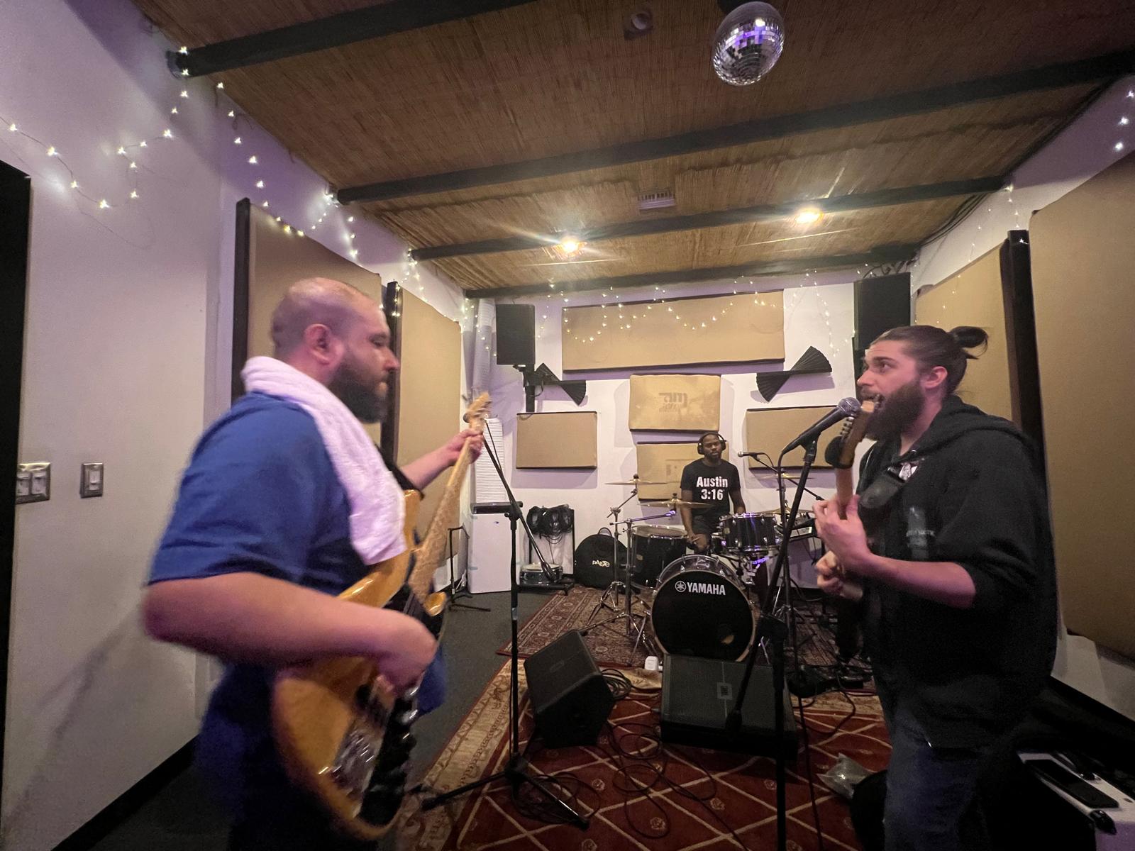 Two men with musical instruments in a recording studio, one playing guitar and the other singing, with a drummer in the background.