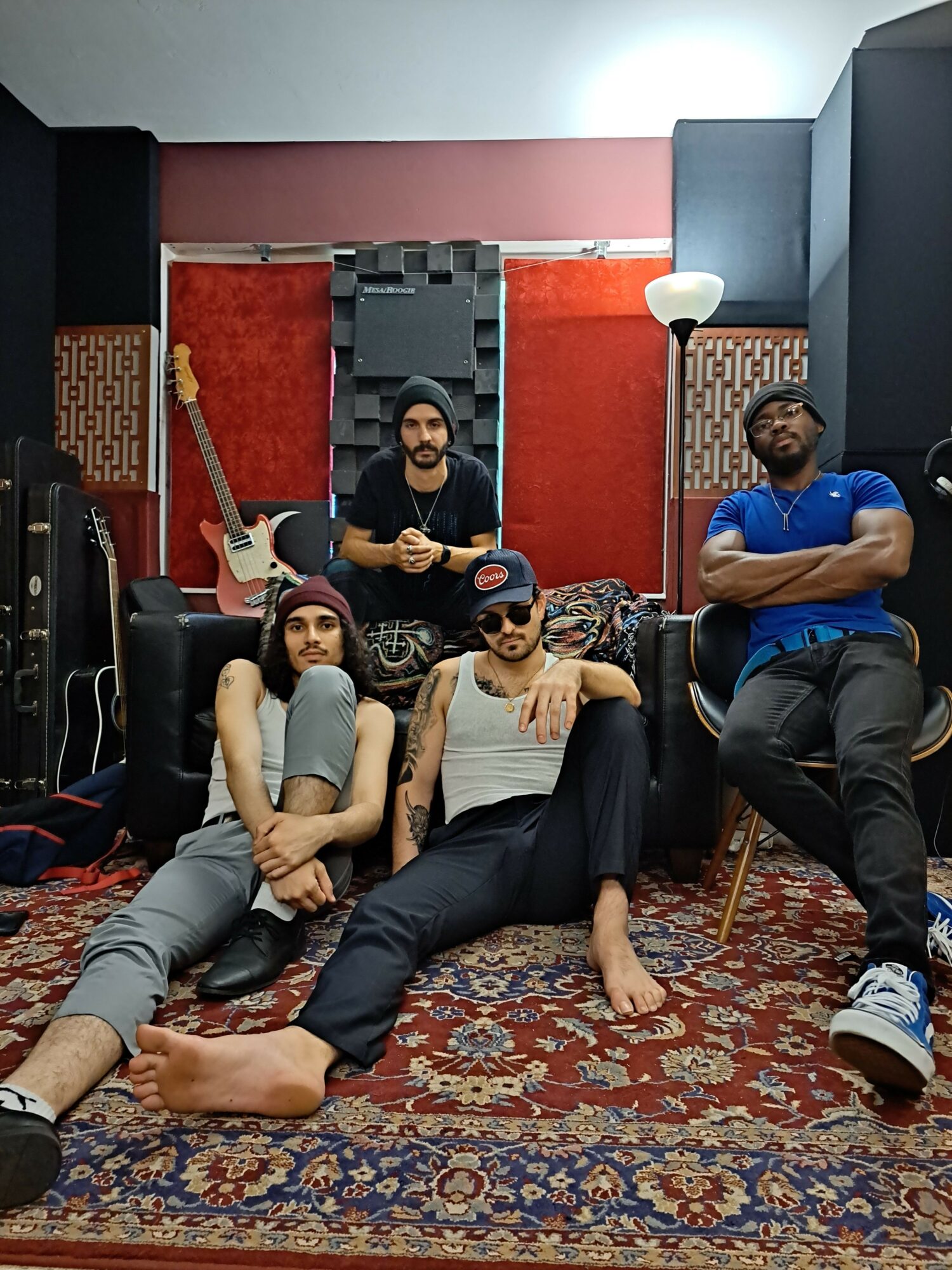Four people in a room with a red wall, sitting and lying on a patterned carpet, some holding props, casual pose.