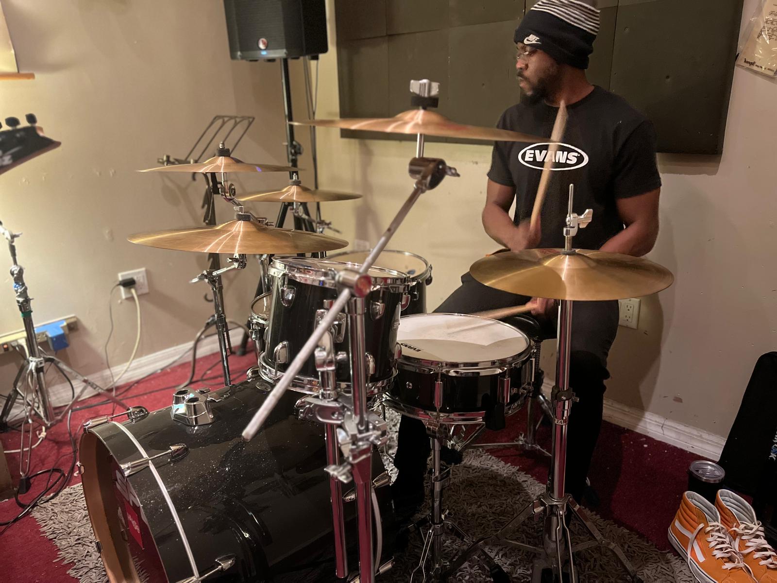 Person wearing a striped beanie and glasses playing a drum set in a room with a TV, carpet, and shoes nearby.