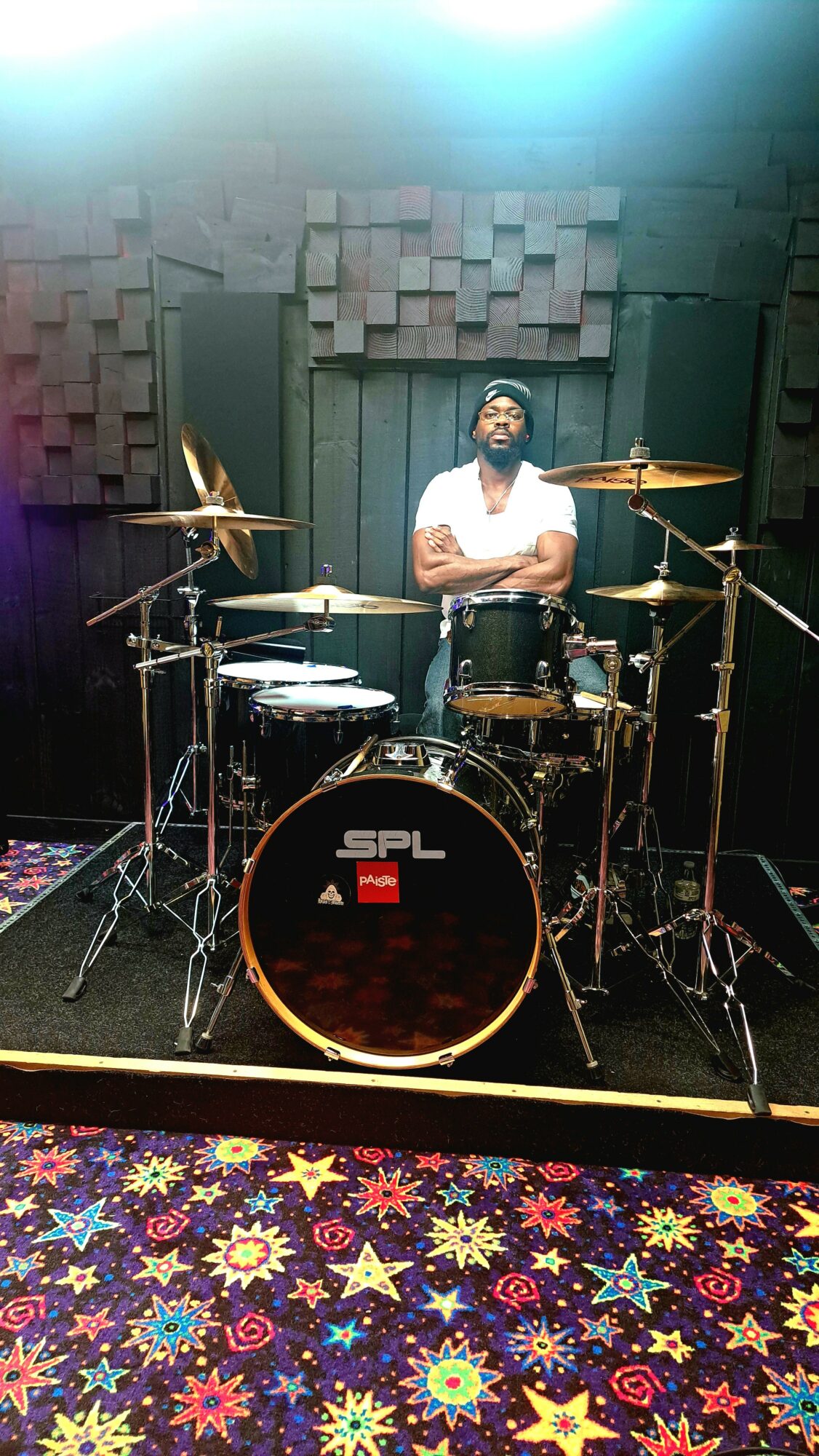 Person standing behind a drum set in a music studio with soundproofing panels, colorful carpet, and bright lighting.