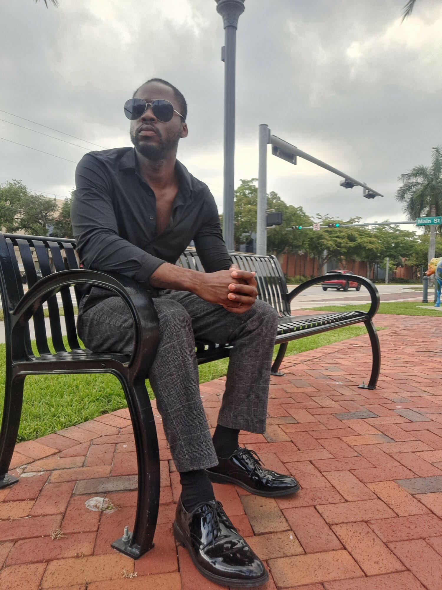 Man sitting on a black chair outdoors on a brick patio, with a park and cloudy sky in the background.