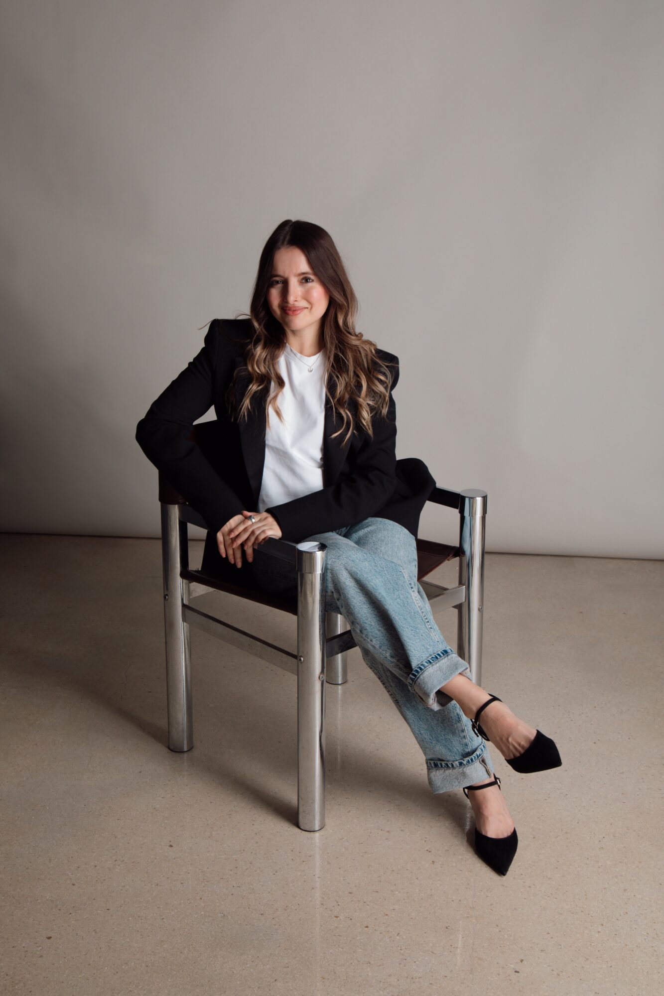 Woman sitting on a chair with crossed legs, wearing a black blazer, white shirt, jeans, and black heels, against a plain background.